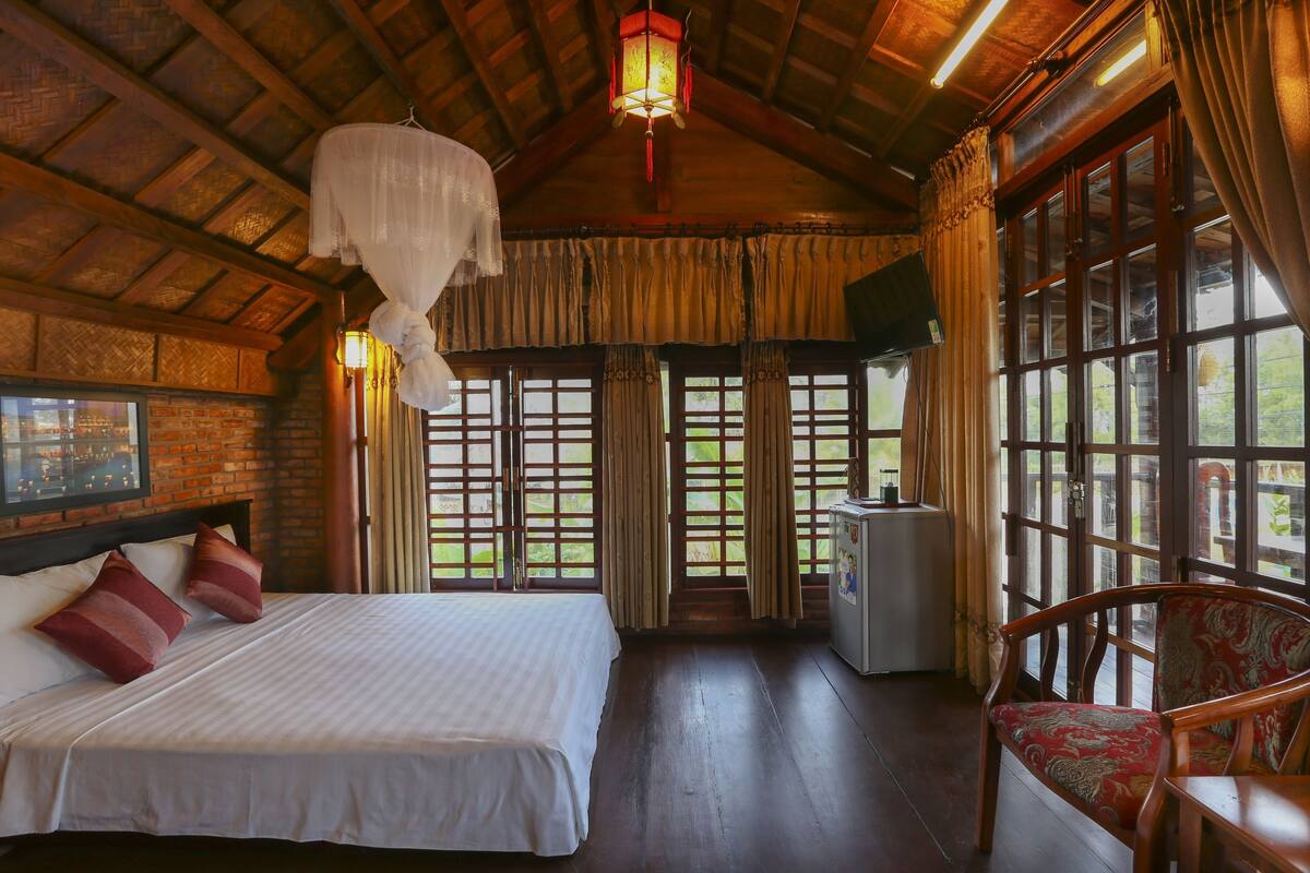 A bedroom with a large bed, wooden ceiling, and a view of greenery outside.