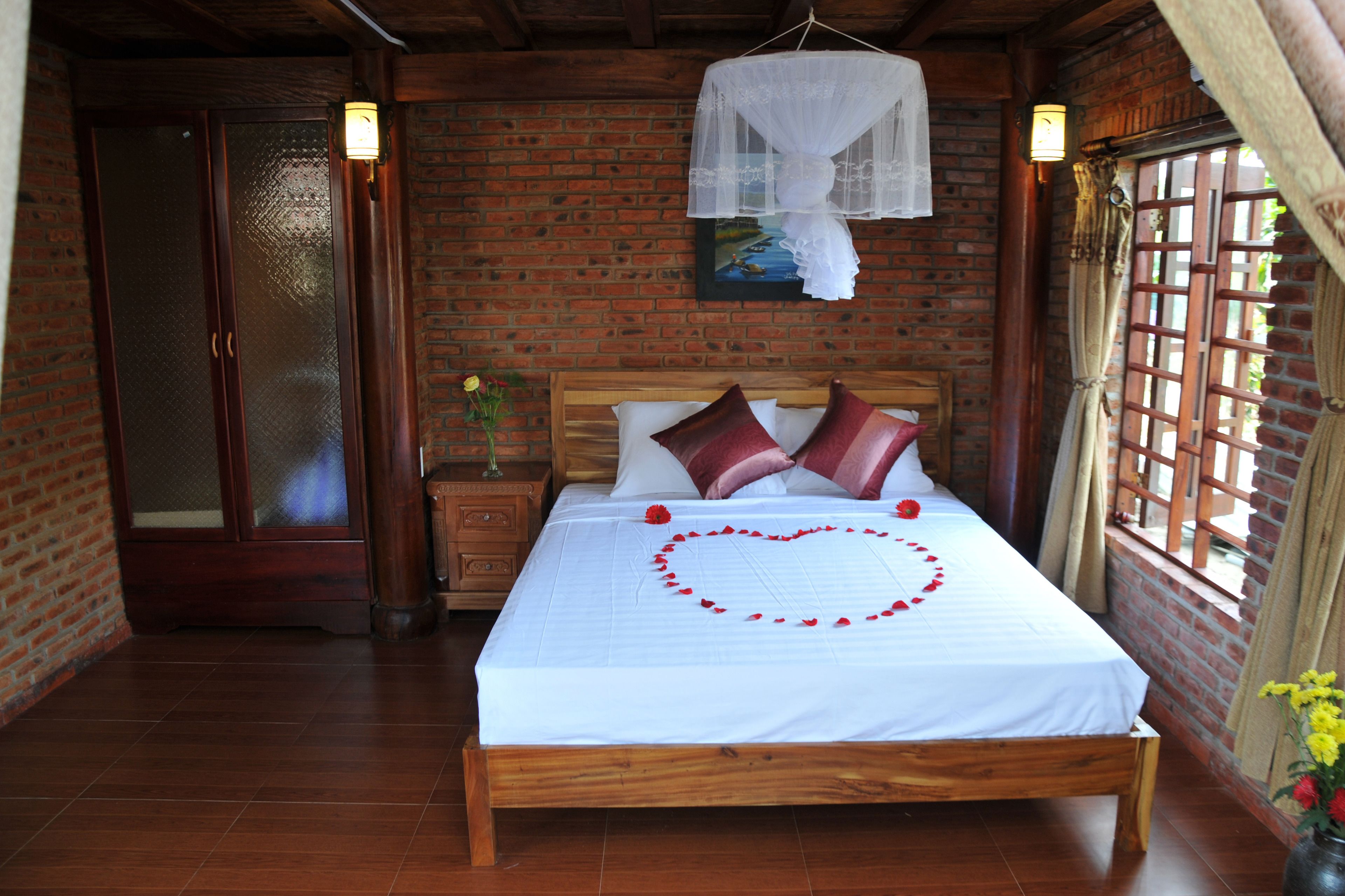 A wooden bed with a heart-shaped decoration of red petals, a white bedspread, and two red pillows.