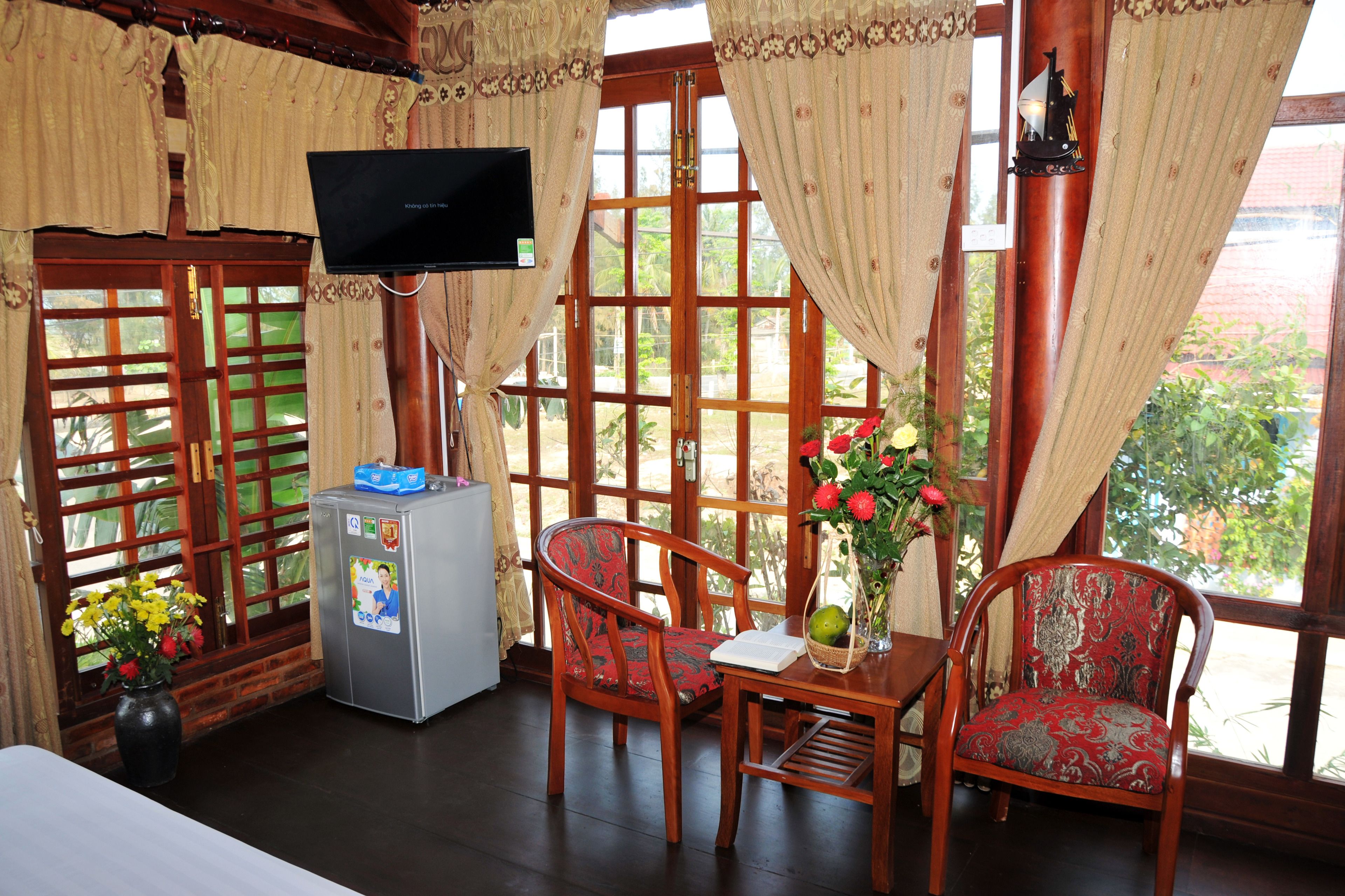 A room with a wooden TV stand, a small refrigerator, two patterned chairs, a table with a vase of flowers, and large windows with curtains.
