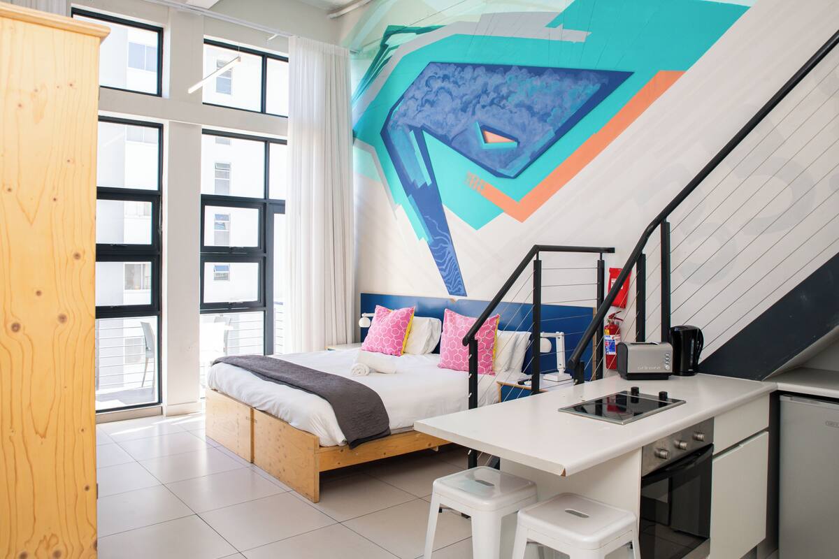 A modern bedroom with a bed, a desk, and a kitchen area.