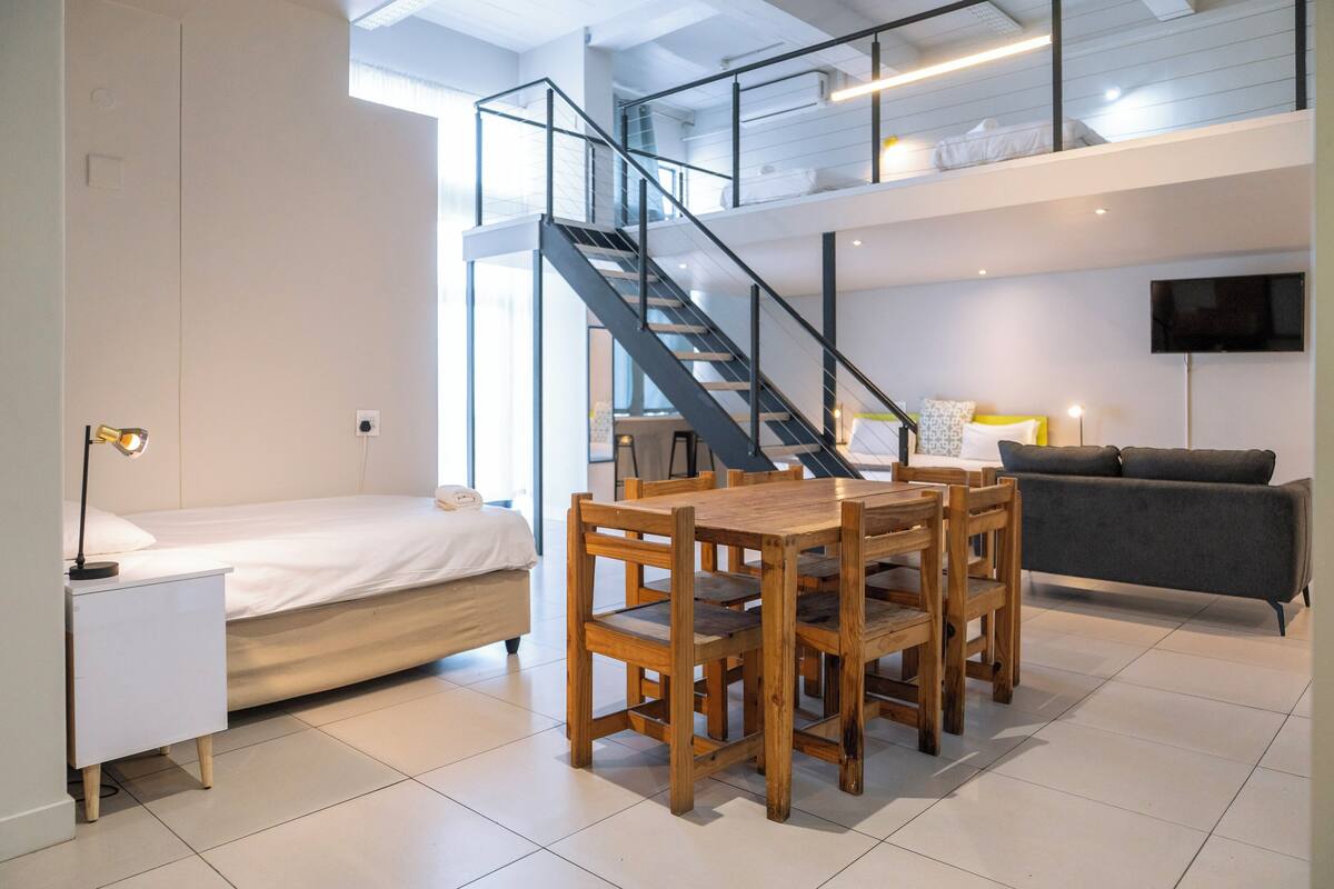 A modern loft-style bedroom with a bed, a wooden dining table, and a staircase leading to an upper level.