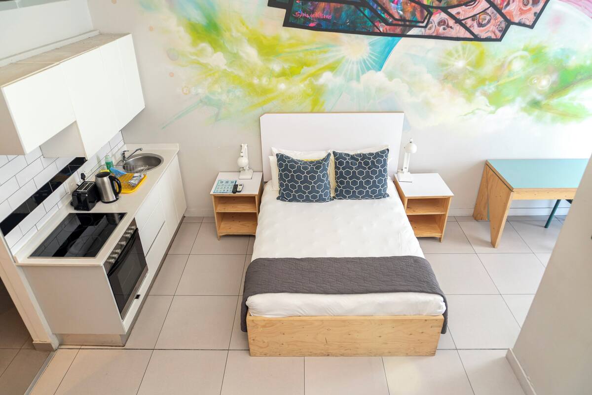 A modern bedroom with a bed, bedside tables, a small wooden desk, and a kitchen area with a stove and sink.