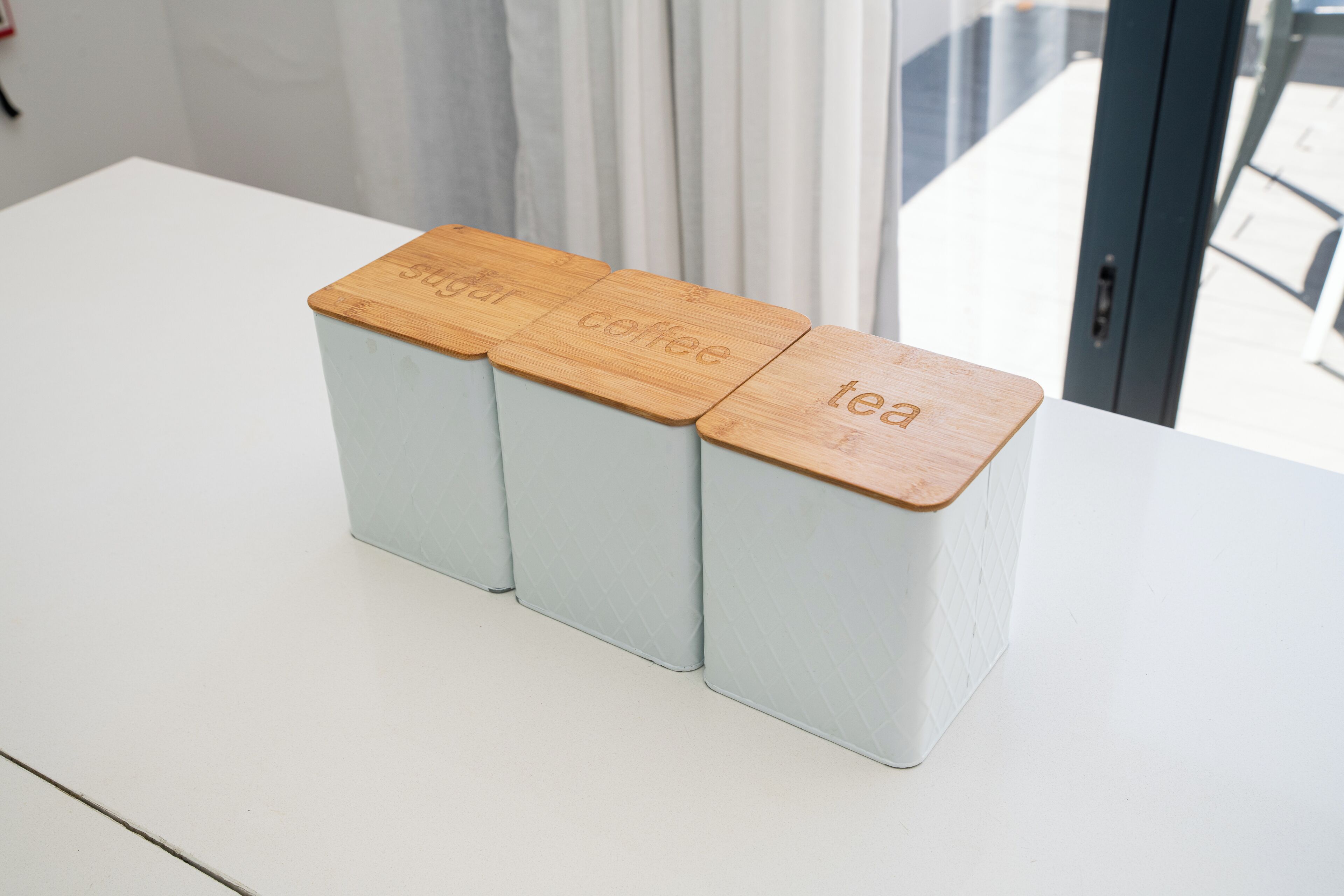 Tres contenedores de almacenamiento con tapas de madera etiquetados como 'azúcar', 'café' y 'té' sobre una mesa blanca.