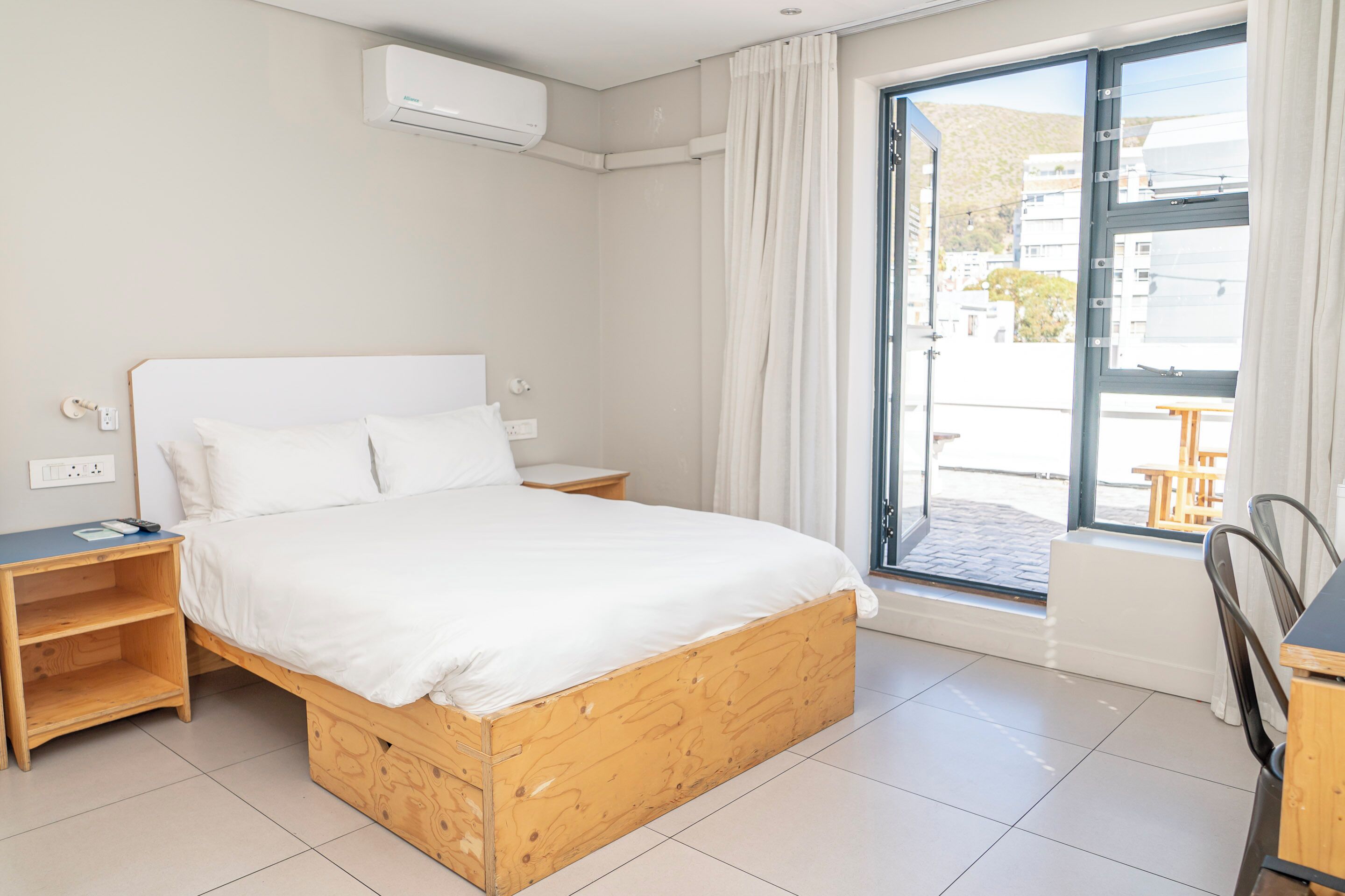 A bedroom with a wooden bed, a nightstand, a desk, and a window with a view of buildings.