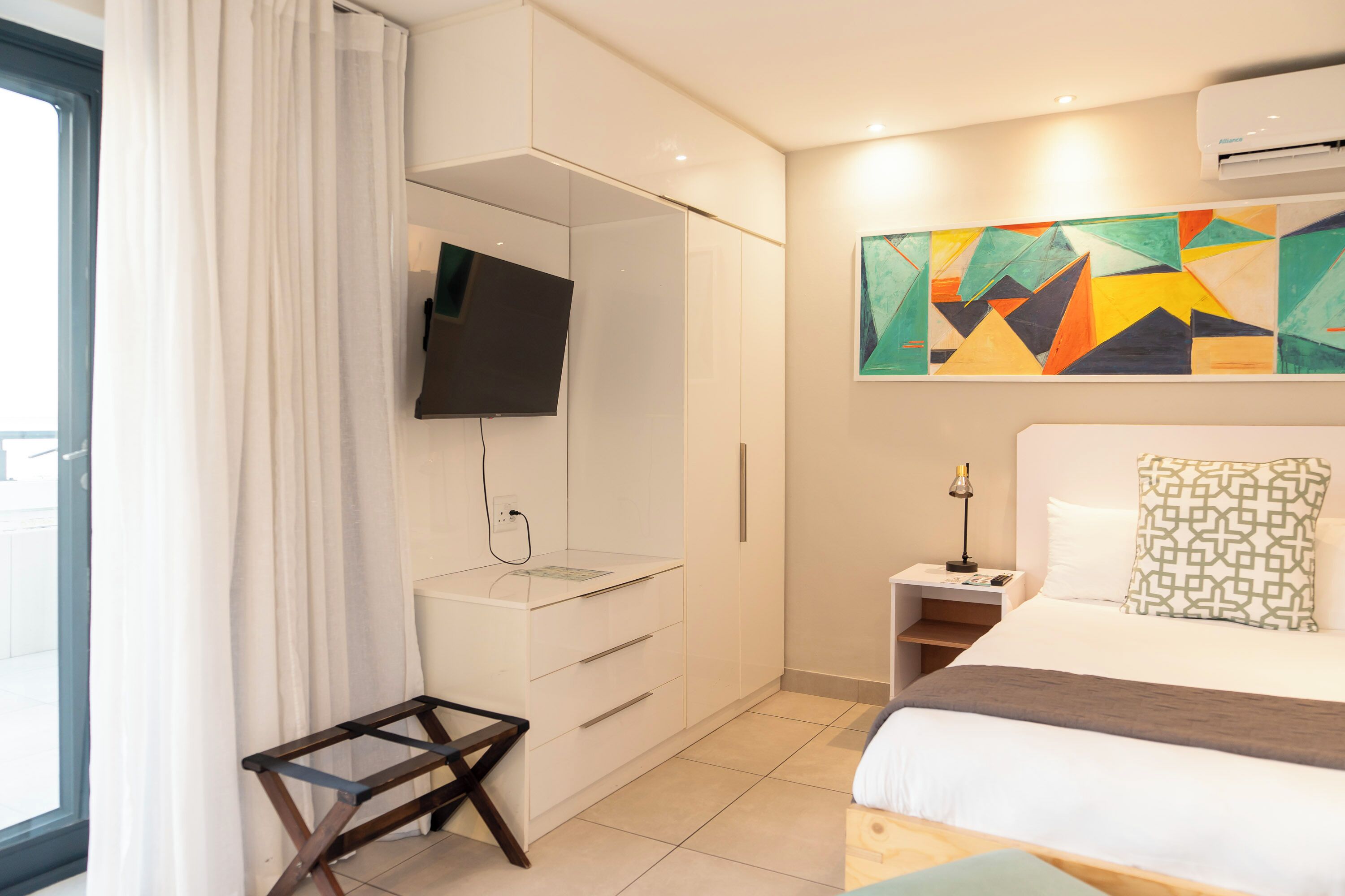 Une chambre d’hôtel moderne, équipée d’un lit, d’un bureau, d’une télévision et ornée d’un tableau abstrait aux couleurs vives.