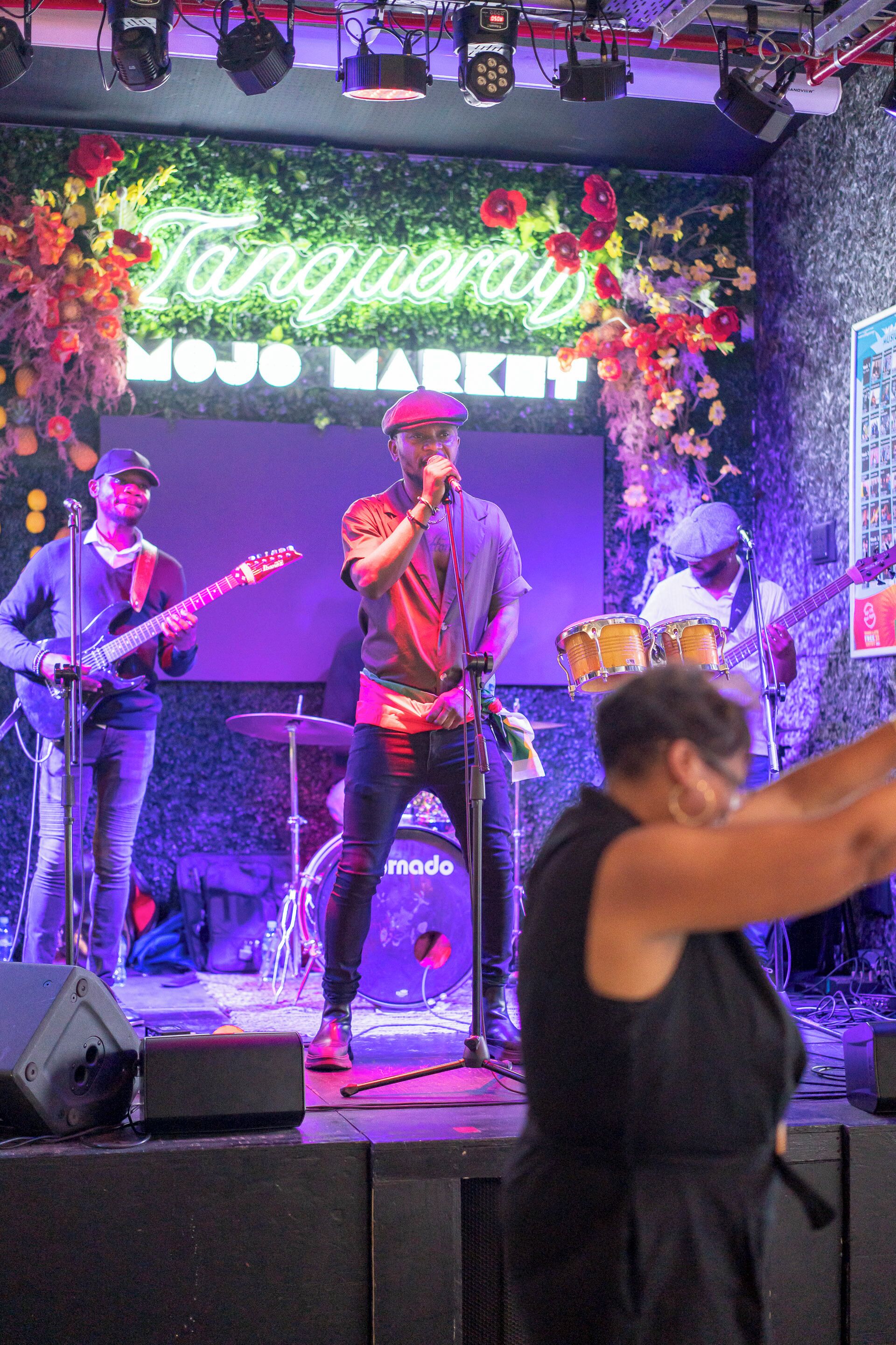 A band performing on a stage with a backdrop that includes the text "Tanqueray" and "MOJO MARKET".