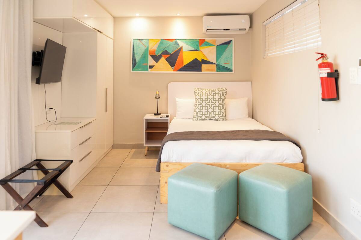 A modern hotel room with a bed, a TV mounted on a cabinet, a colorful abstract painting on the wall, and a fire extinguisher.