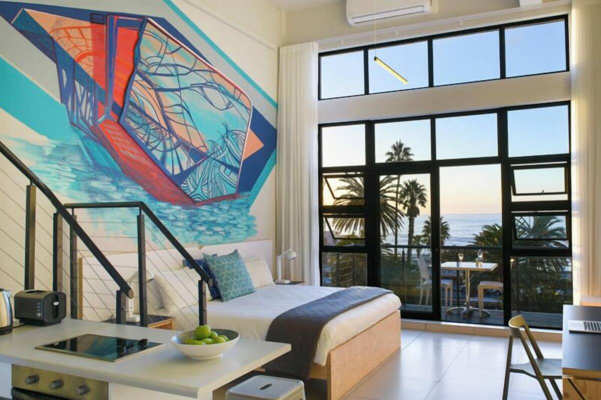 A modern hotel room with a large window, a bed, a desk with a laptop, a coffee maker, and a painting on the wall.