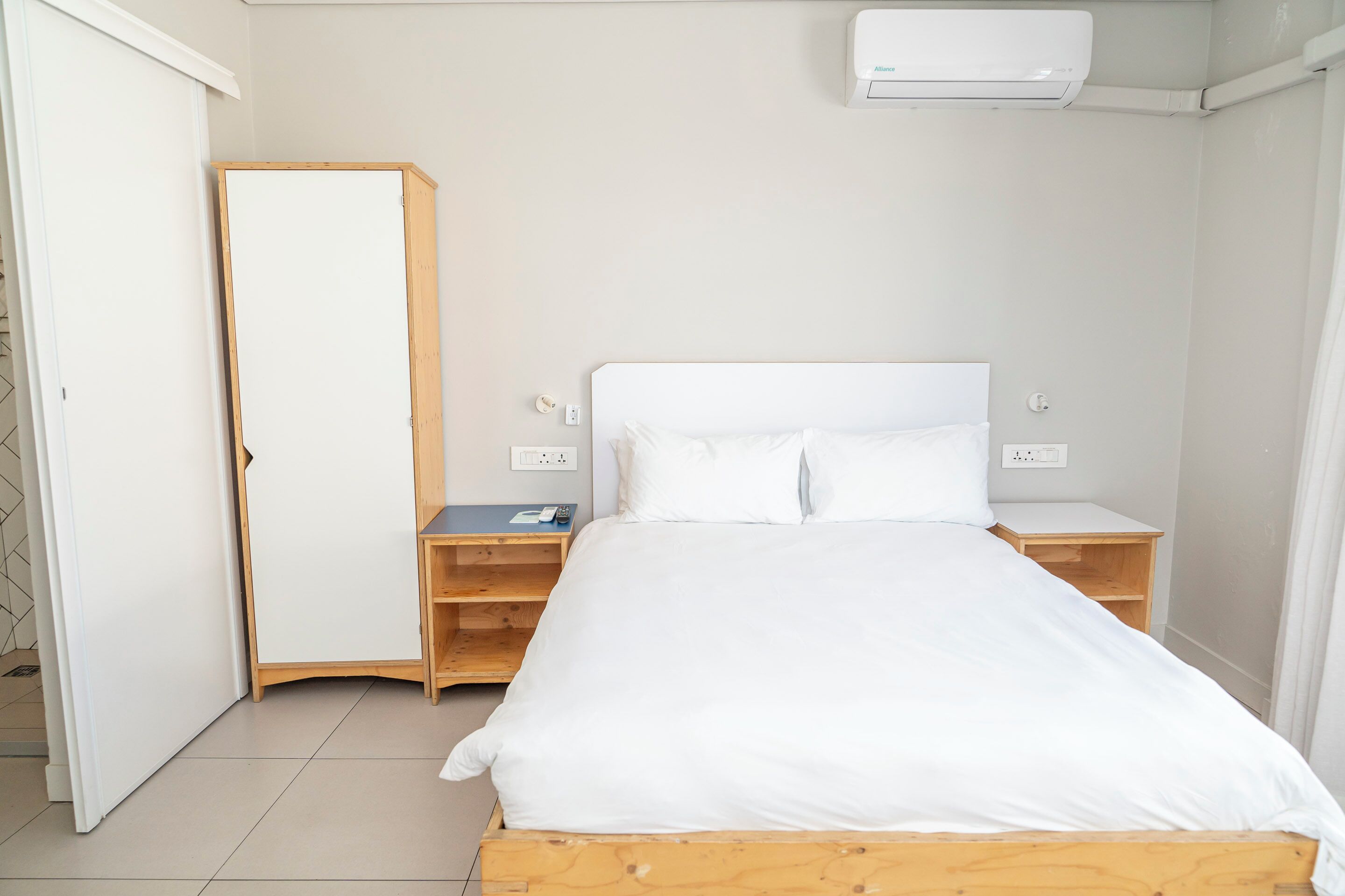 A bedroom with a bed, wooden nightstands, a wardrobe, and an air conditioning unit.