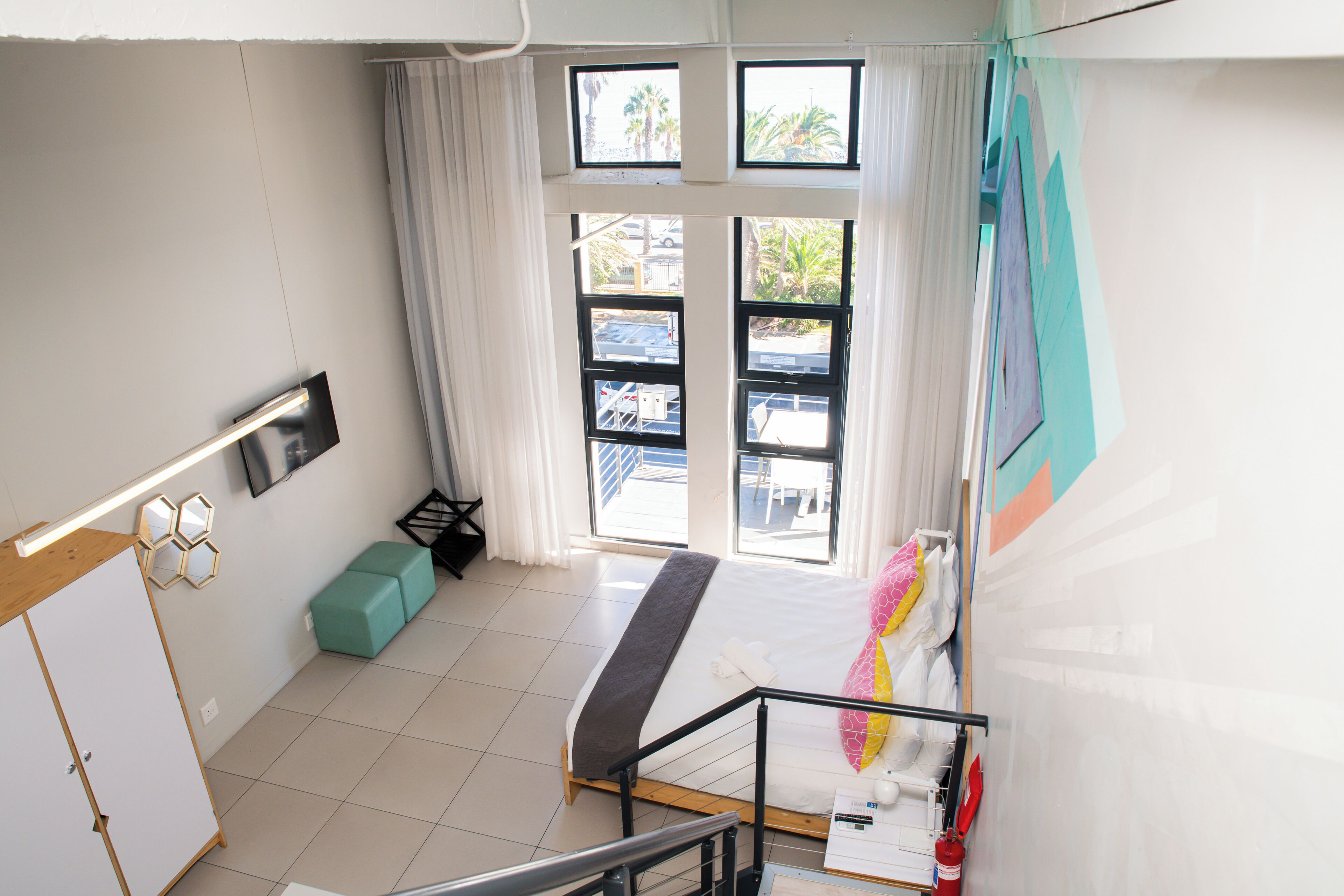 Habitación de hotel con cama, escritorio, silla, ventana con cortinas, extintor y vistas al exterior.