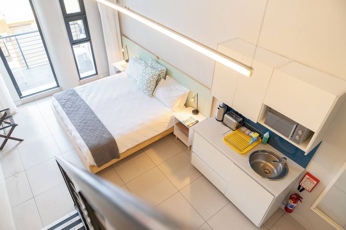A modern hotel room with a bed, bedside table, lamp, and a small kitchenette with a sink and microwave.