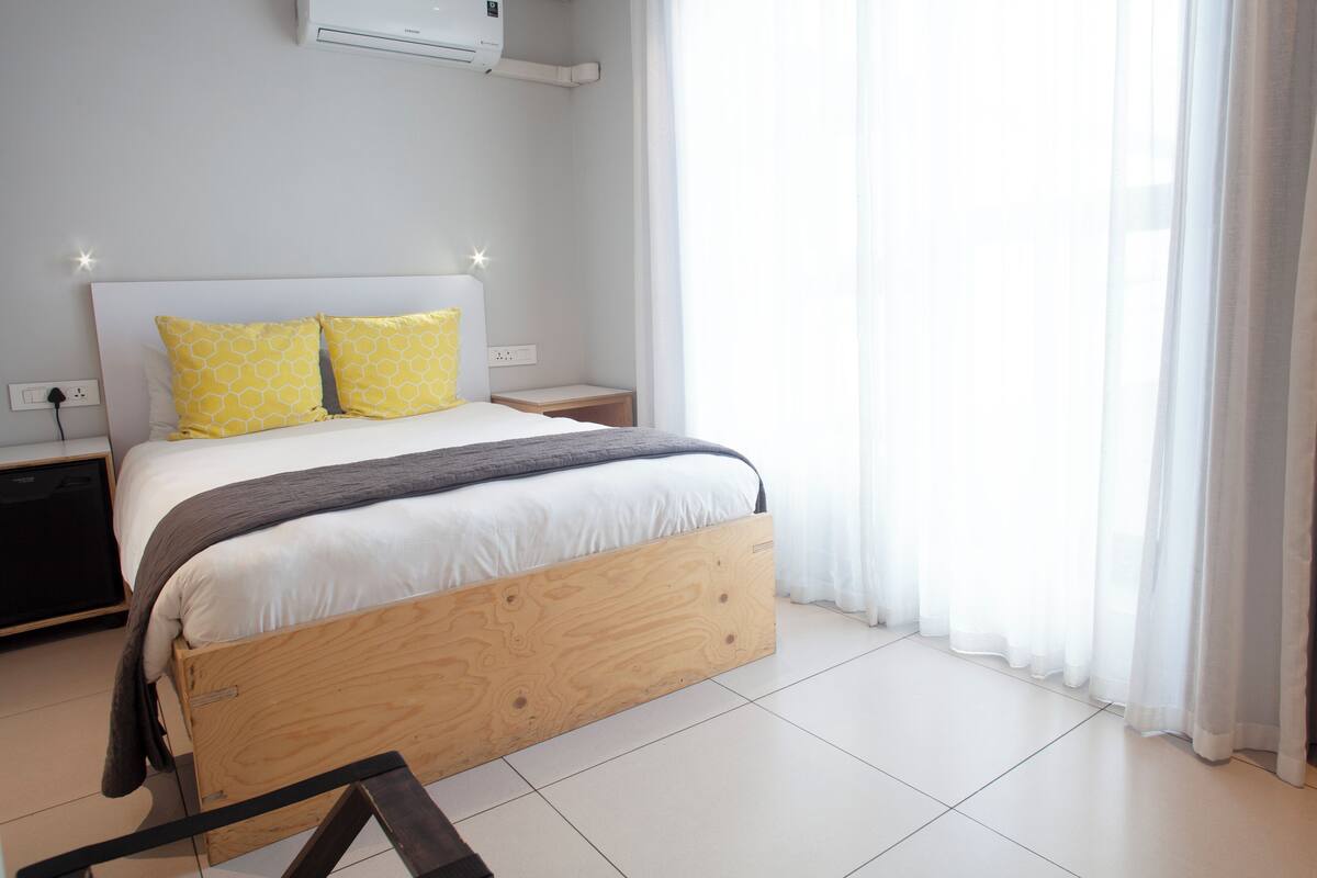 A neatly made bed with a wooden frame, two yellow patterned pillows, and a white comforter. There is a bedside table with a lamp, a power outlet, and a small wooden nightstand. The room has white walls, a tiled floor, and a large window with sheer curtains.