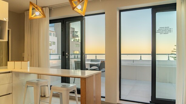 A modern kitchen with a bar stool, a wooden table, and a view of the city through large windows. - Mojo Hotel (Cape Town)