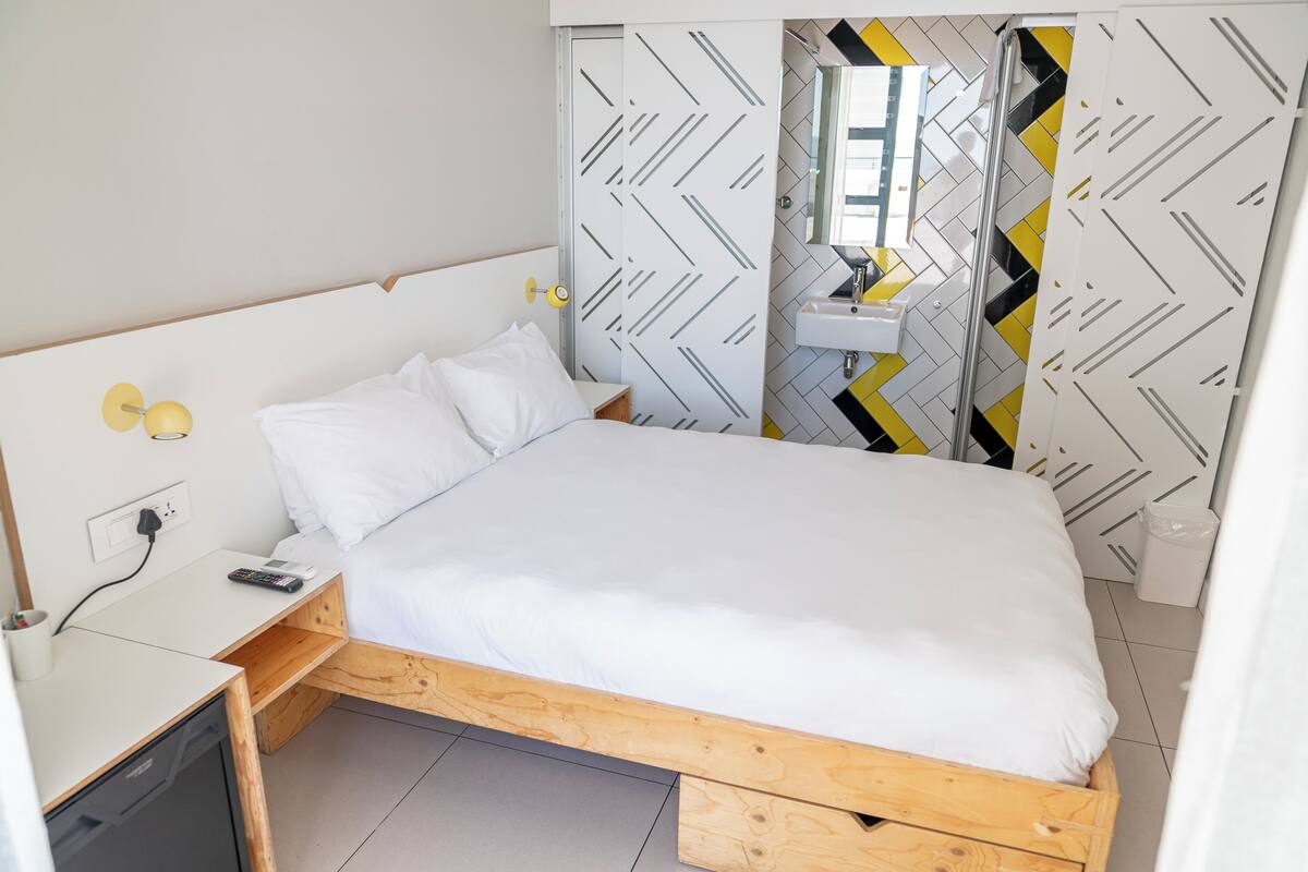 A compact bedroom with a single bed, a bedside table with a phone, and a small desk with a dishwasher.
