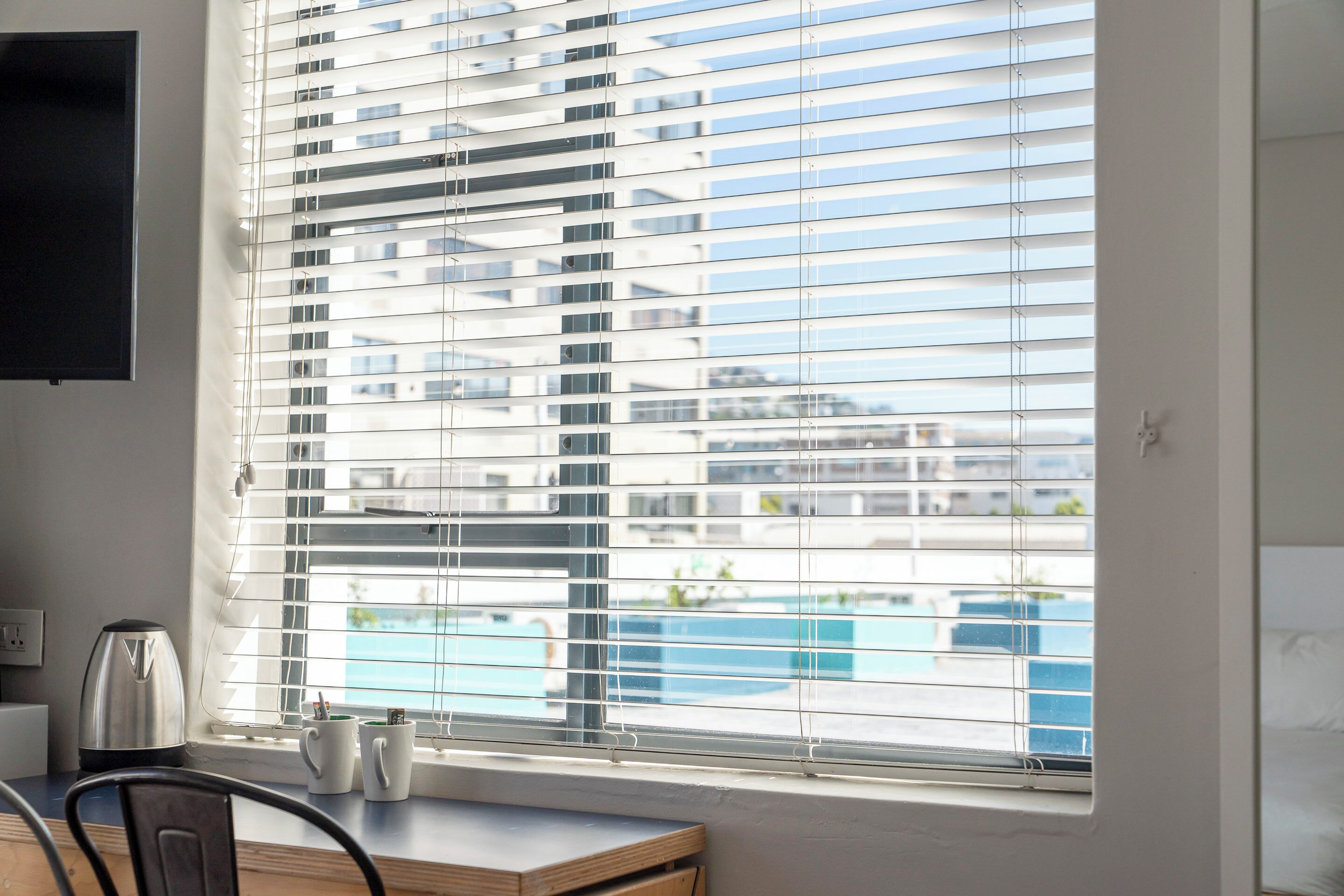 A room with a large window, a flat-screen TV mounted on the wall, a kettle, and a small table with two mugs.
