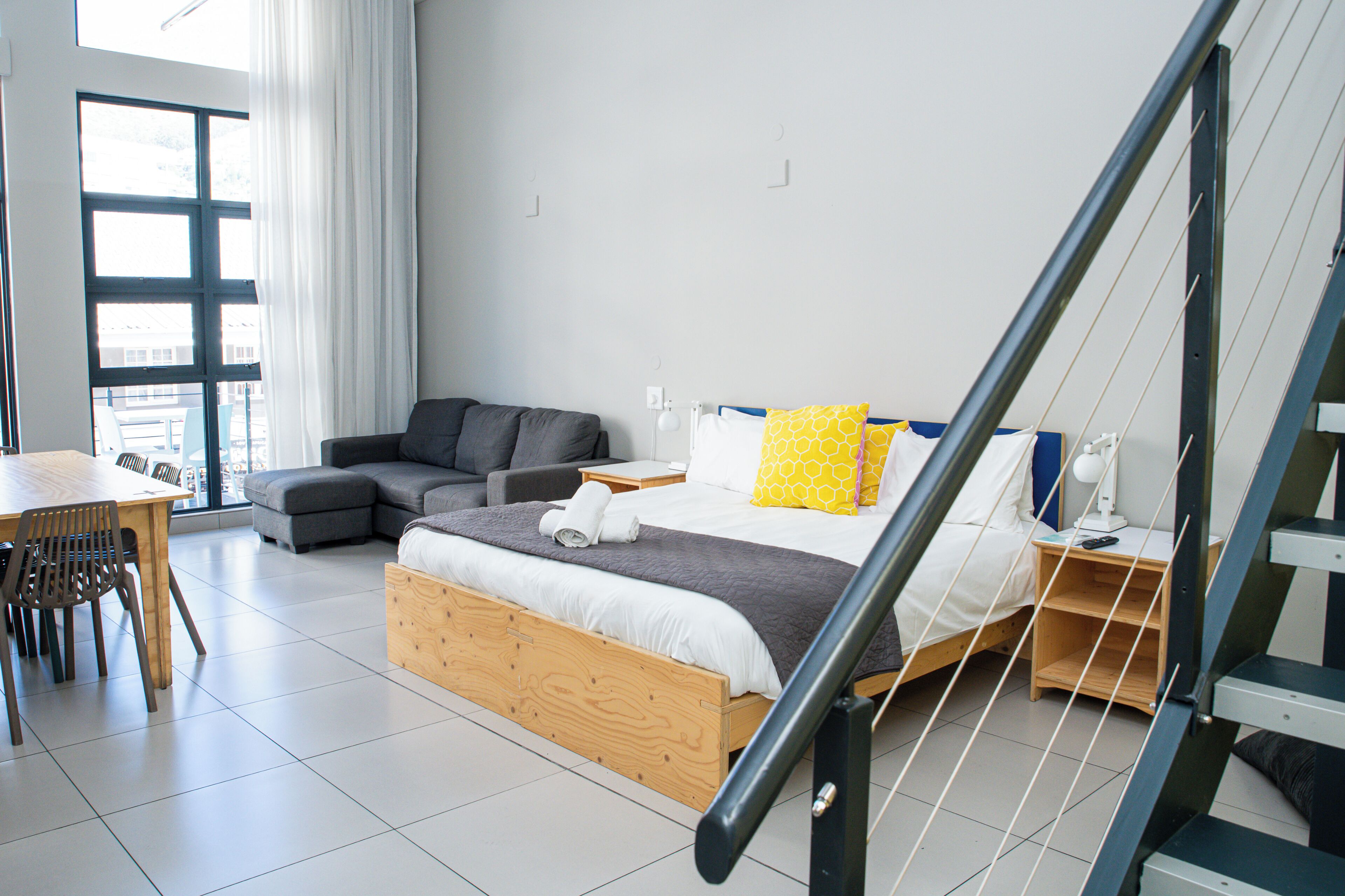 A modern bedroom with a wooden bed, a gray sofa, a dining table, and a staircase.