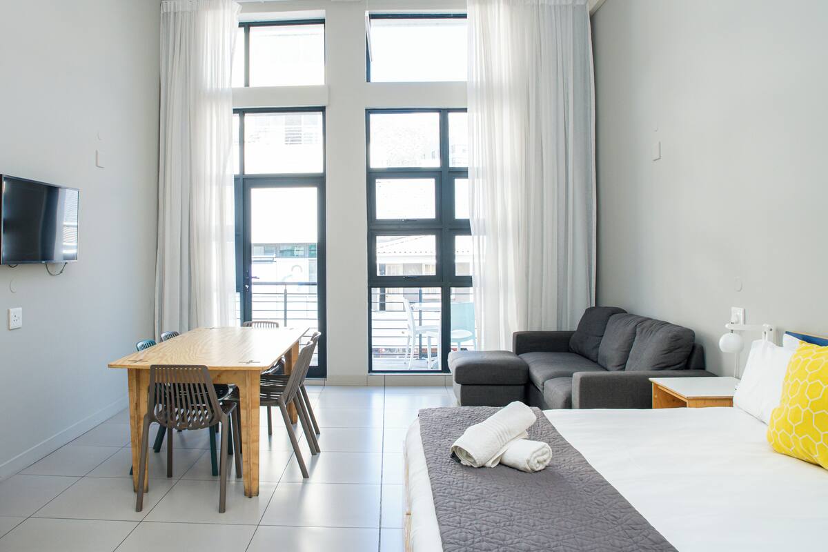 A hotel room with a bed, a sofa, a dining table with chairs, and a TV mounted on the wall.