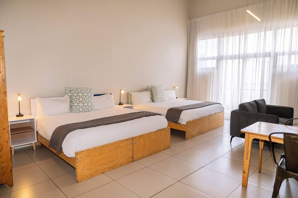 A hotel room with two wooden beds, a wooden table, a chair, and a large window with sheer curtains.