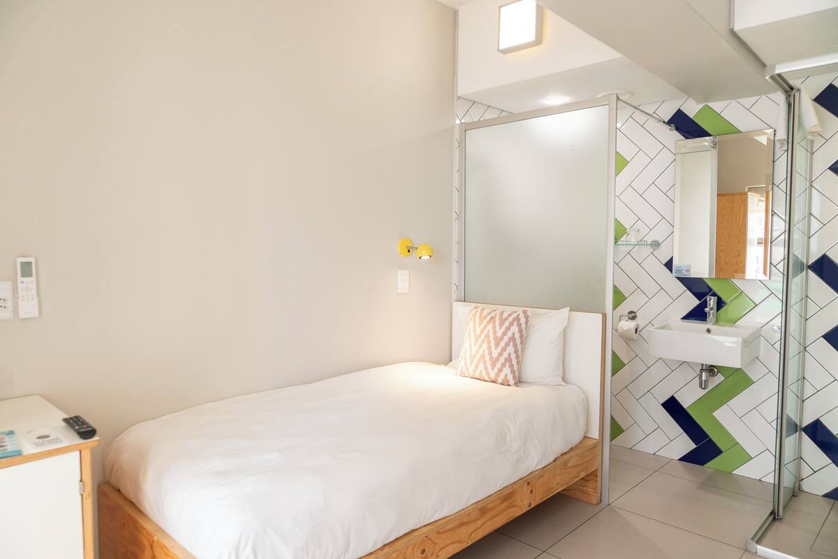 A bedroom with a bed, a bedside table, a mirror, and a bathroom with a sink and shower.