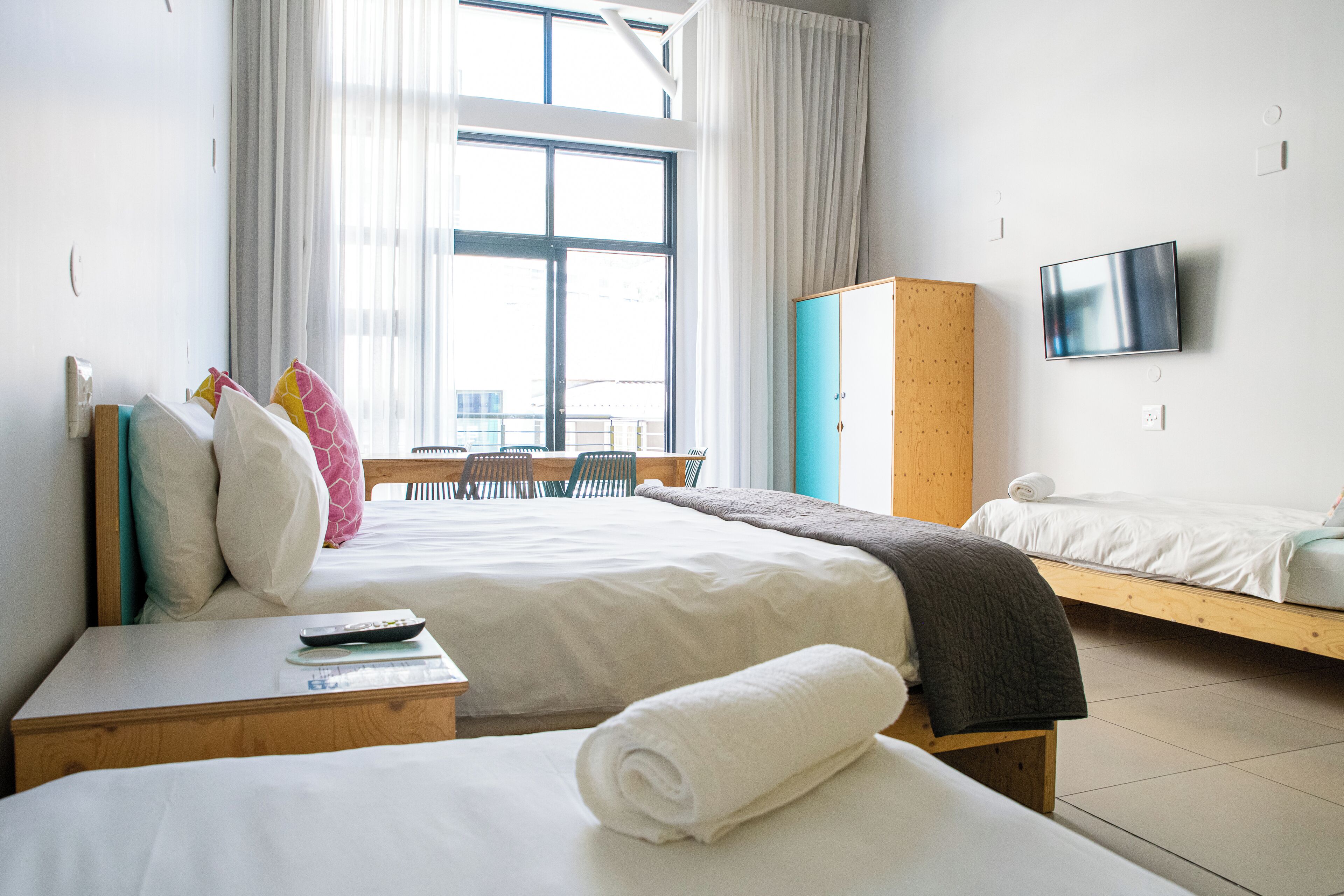 A hotel room with two beds, a TV mounted on the wall, a wooden wardrobe, and a window with curtains.