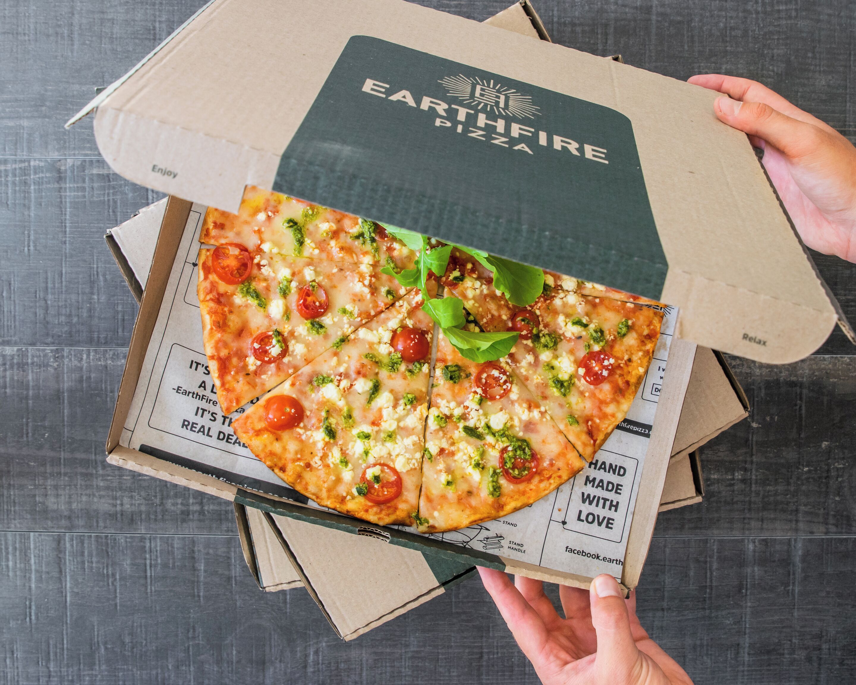A hand holding a pizza box with a slice of pizza visible.