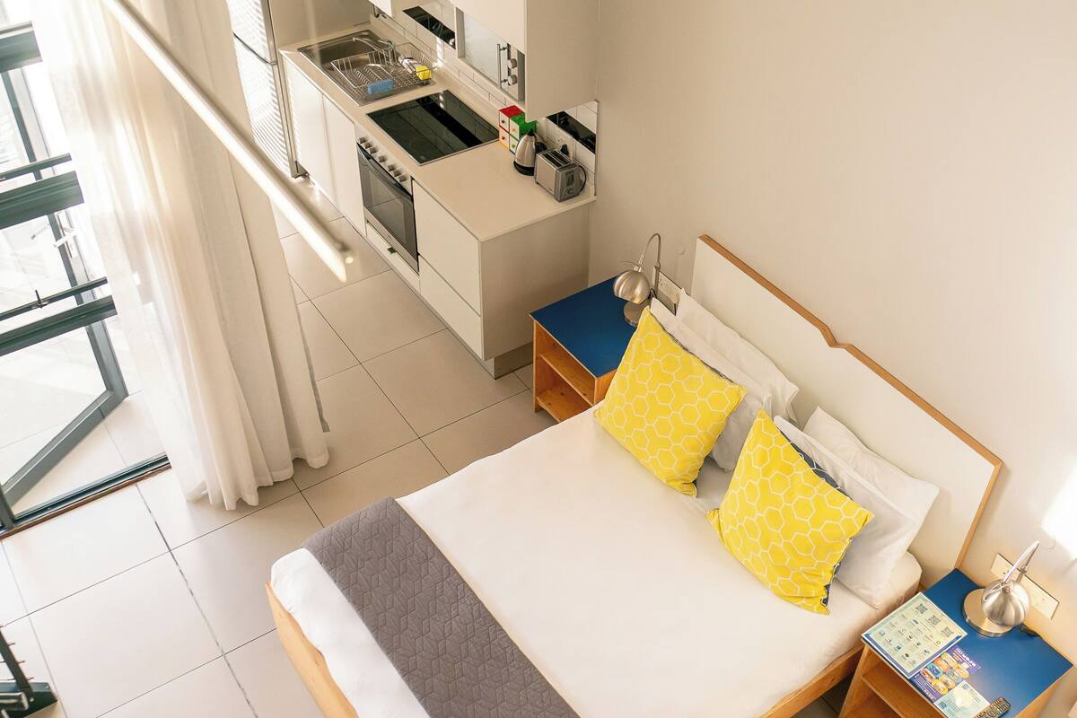A modern hotel room with a large bed, a small kitchenette, and a balcony with curtains.