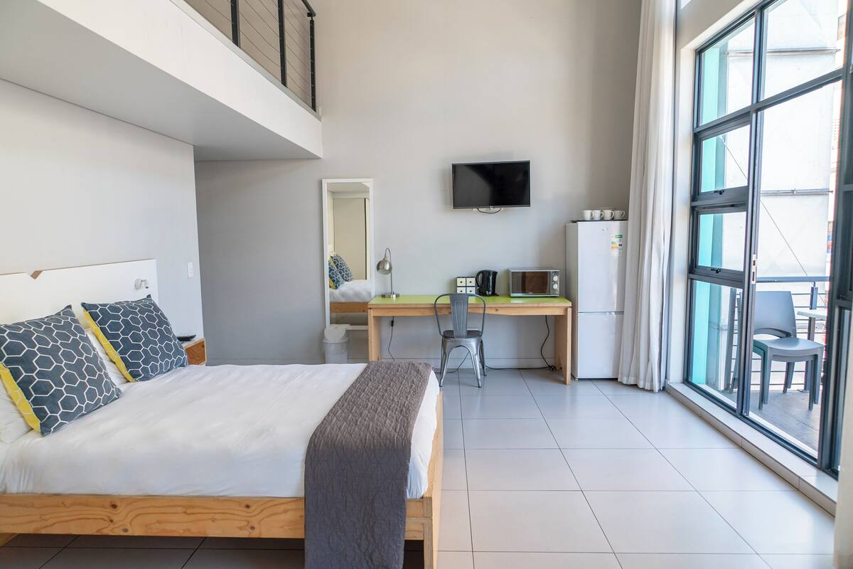 A modern hotel room with a bed, desk, chair, TV, and large windows.