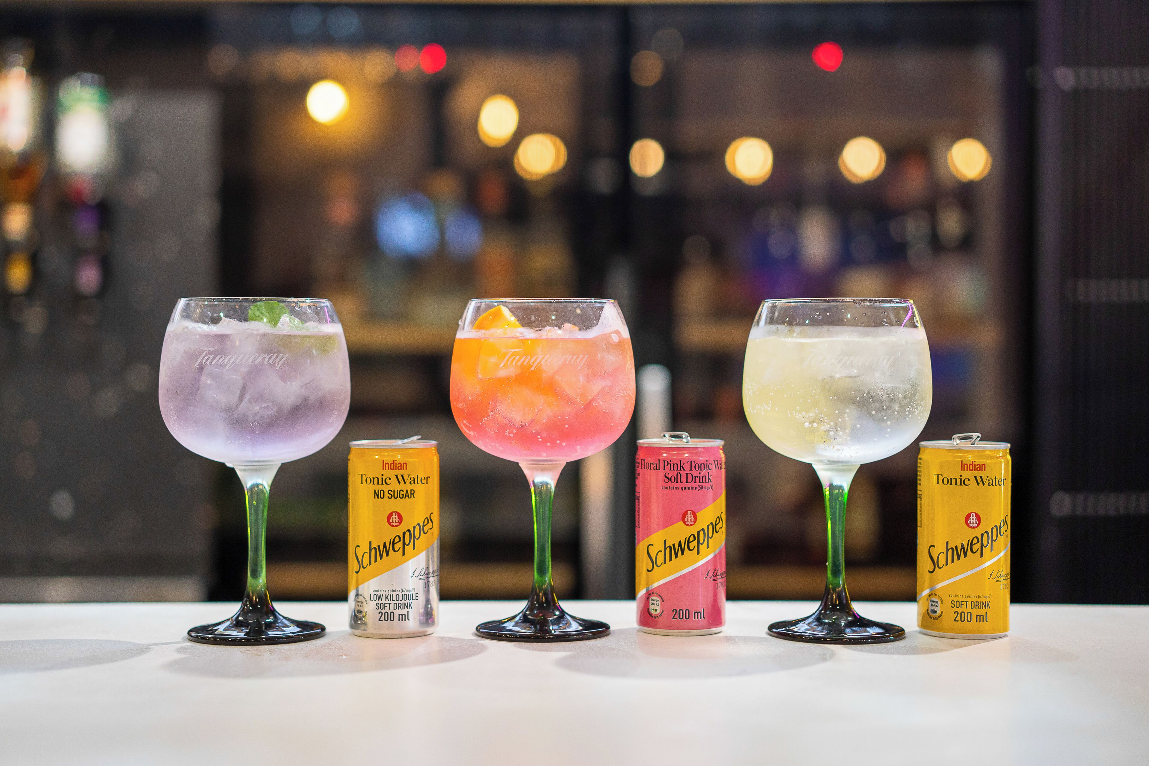 Trois verres de cocktails à base d’eau tonique, accompagnés de canettes de Schweppes.