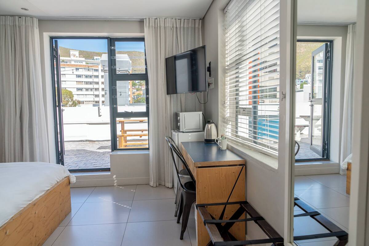 A hotel room with a bed, a desk with a TV, a kettle, and a view of the outside balcony.