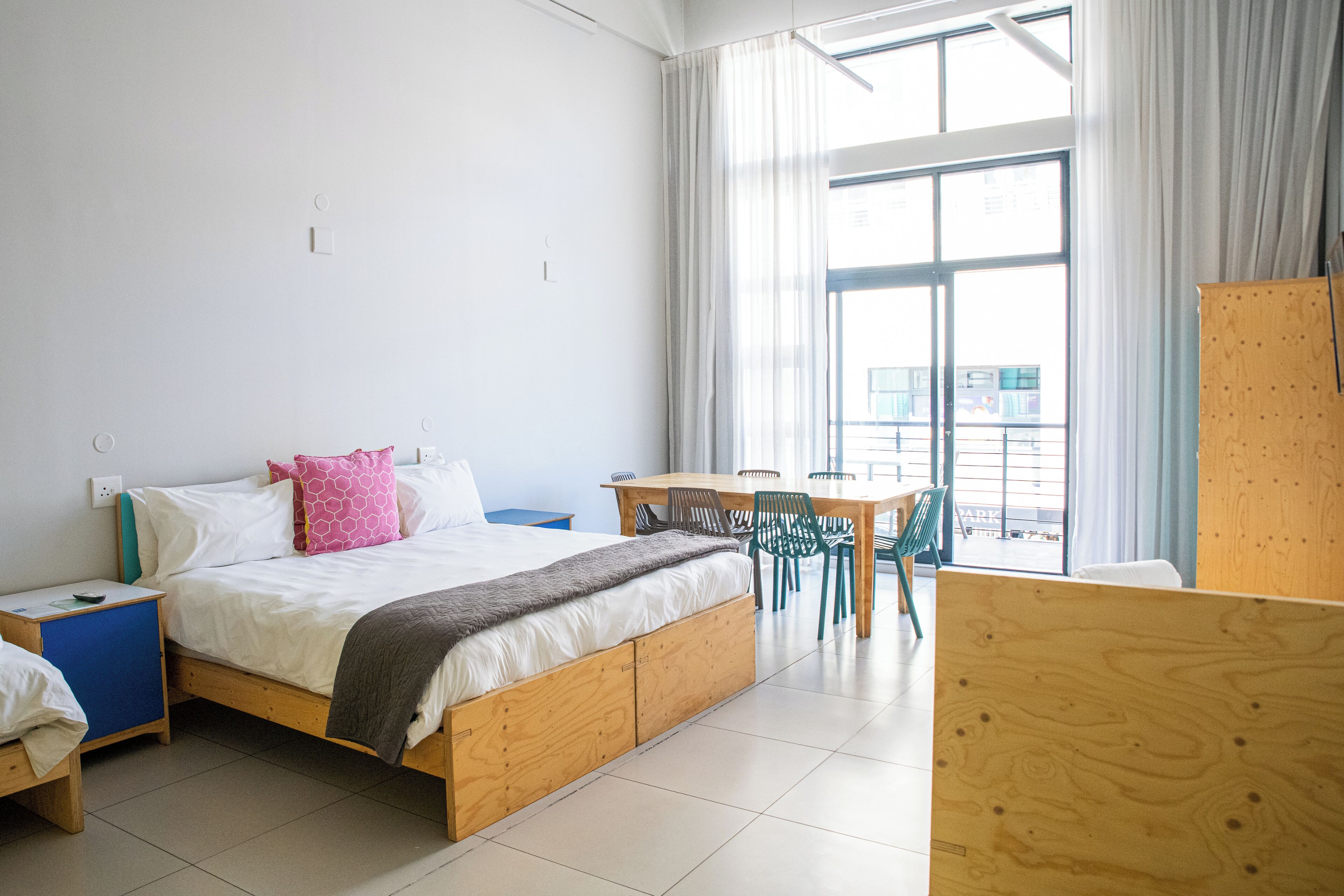 Camera da letto moderna con un letto di legno, una scrivania e sedie, ampie finestre con tende e vista sulla città.