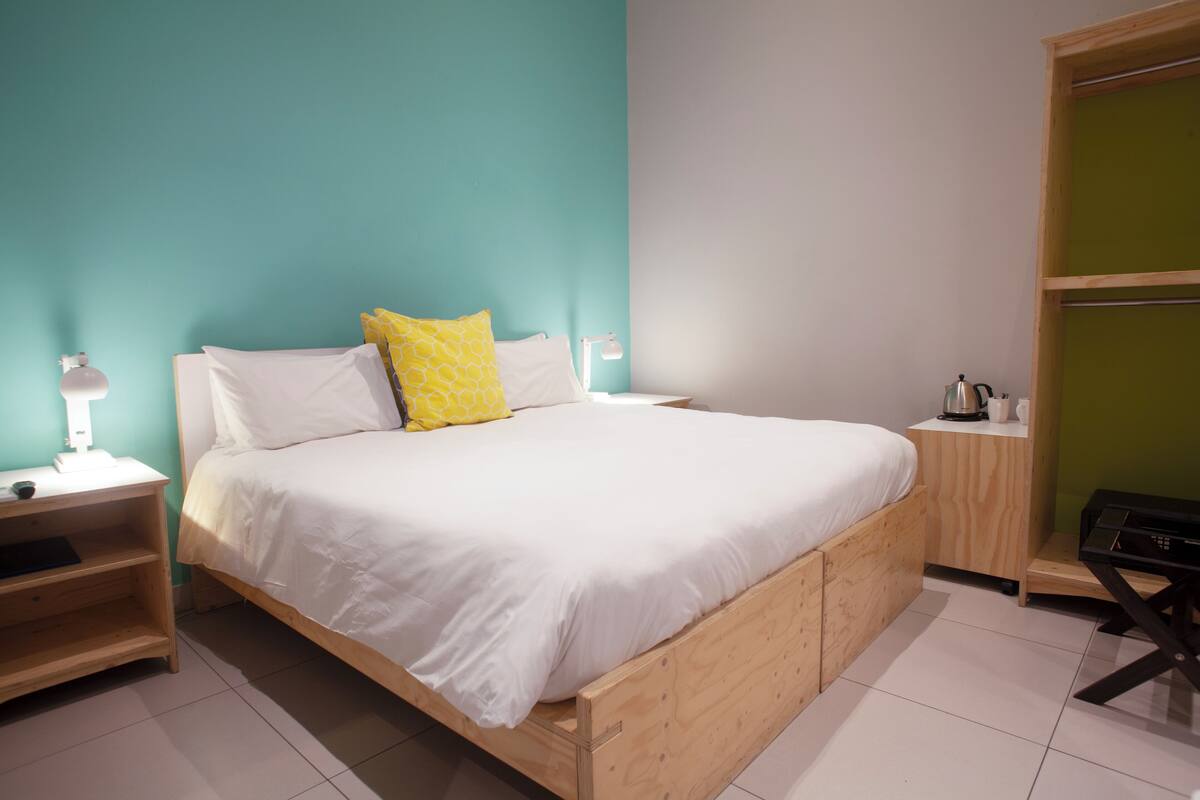 A bedroom with a wooden bed, white bedding, a yellow patterned pillow, a bedside table with a lamp, and a wooden nightstand.