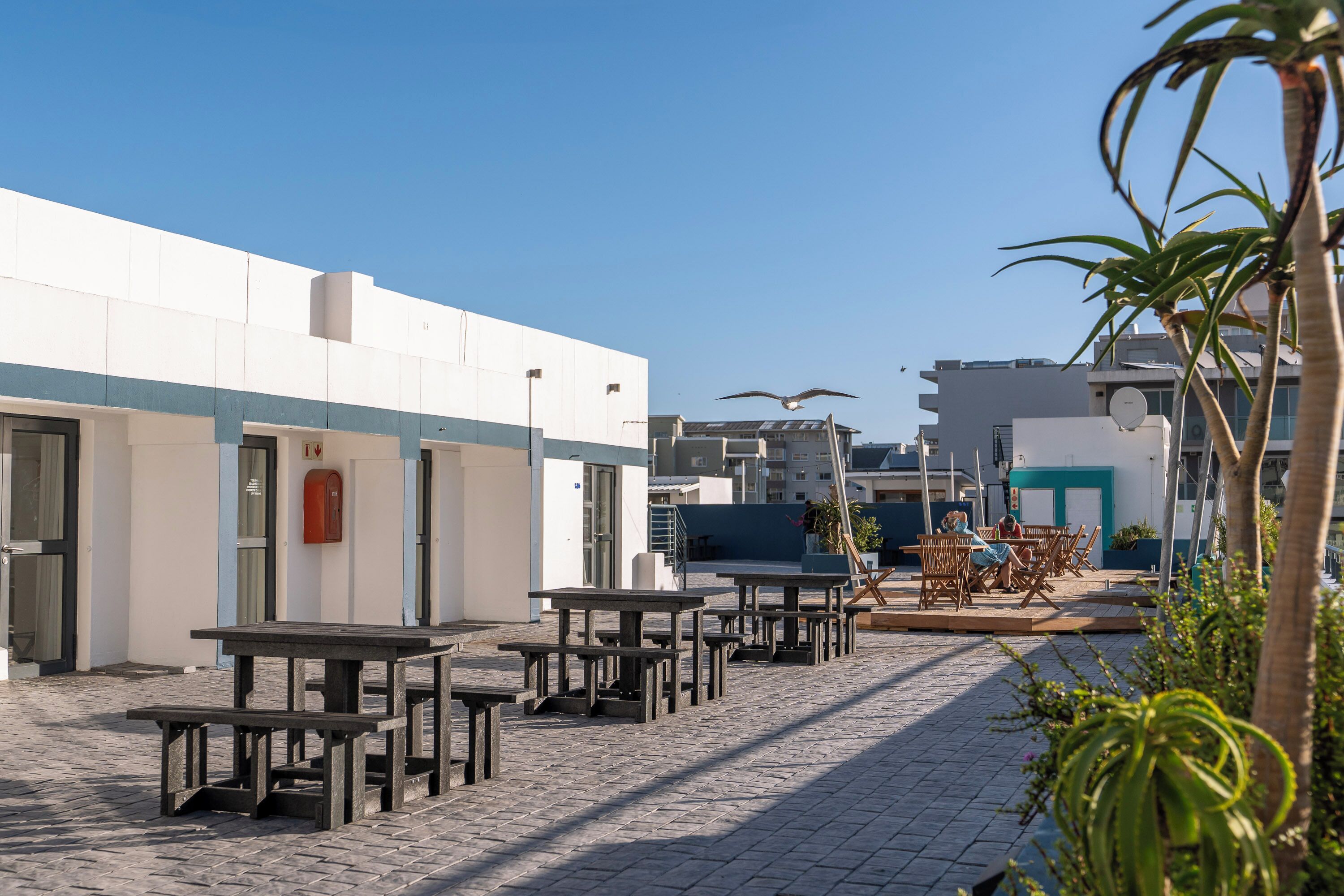 Un patio al aire libre con mesas y sillas, un edificio con puerta azul y cielo despejado.