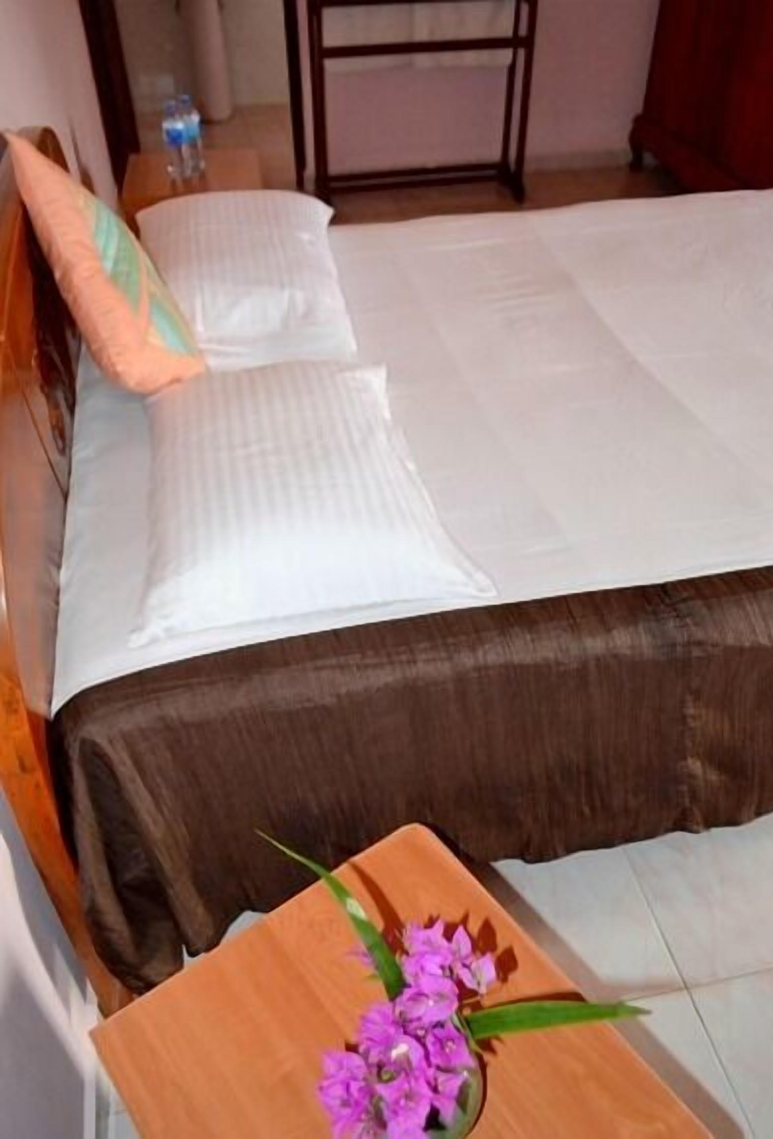A neatly made bed with white linens, a wooden nightstand with a vase of purple flowers, and a water bottle on the floor.