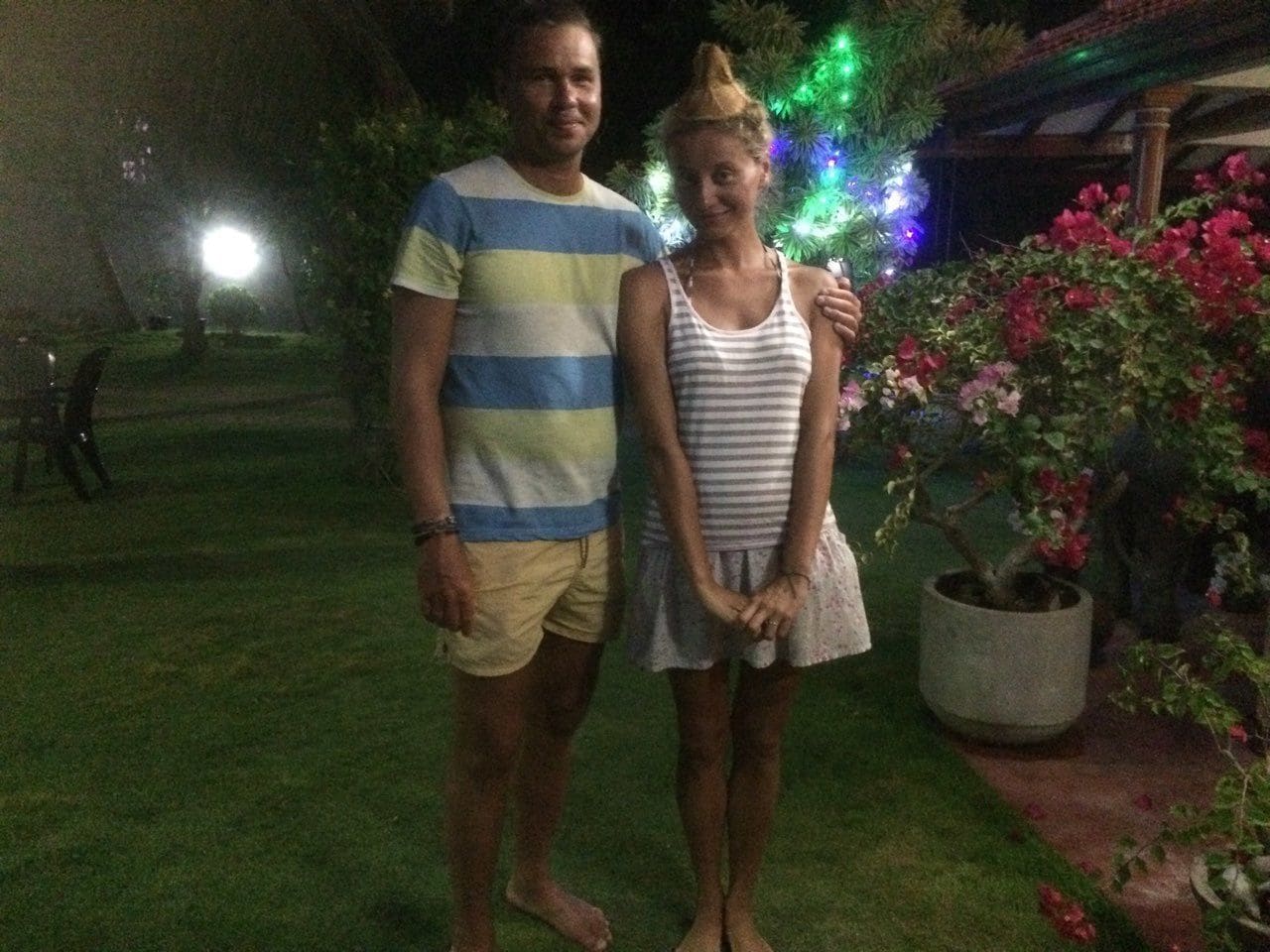 Two people standing in a garden at night with a Christmas tree and potted plants in the background.