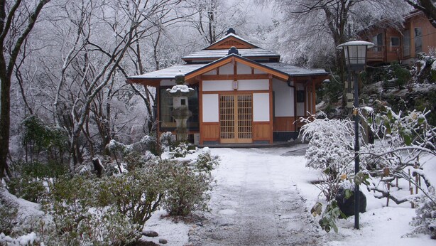 Un edificio japonés tradicional con fachada de madera y tejado de tejas, rodeado de árboles cubiertos de nieve y un sendero nevado que conduce a la entrada.