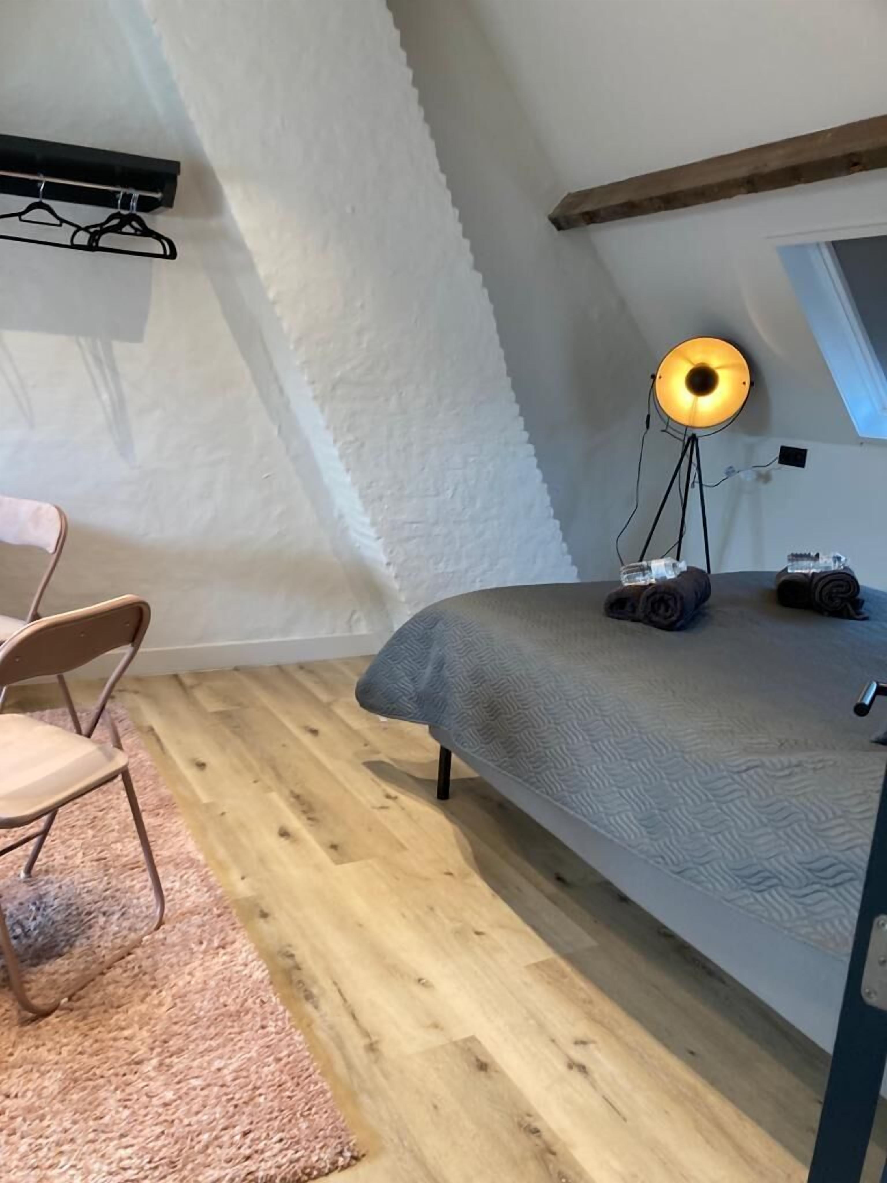 A small, cozy room with a bed, a chair, a floor lamp, and a window.