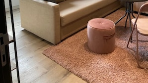 Un ambiente tipo loft con techo alto, tragaluces, un sofá color beige con cojines, un otoman redondo, una mesita redonda y una silla.