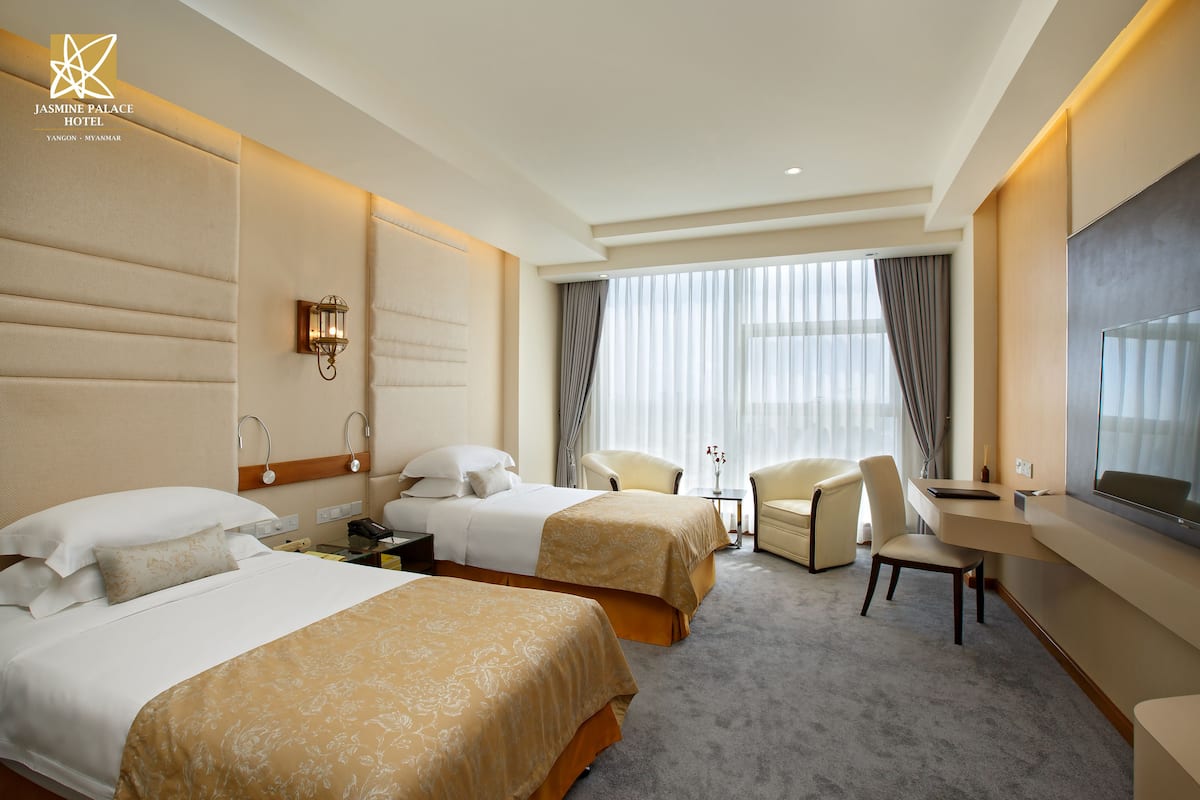 A hotel room with two beds, a desk, a chair, a TV, and a large window with curtains.