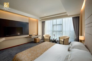 A modern hotel room with a large bed, a flat-screen TV mounted on the wall, two armchairs, a small side table, and a large window with curtains. - Jasmine Palace Hotel (Yangon)