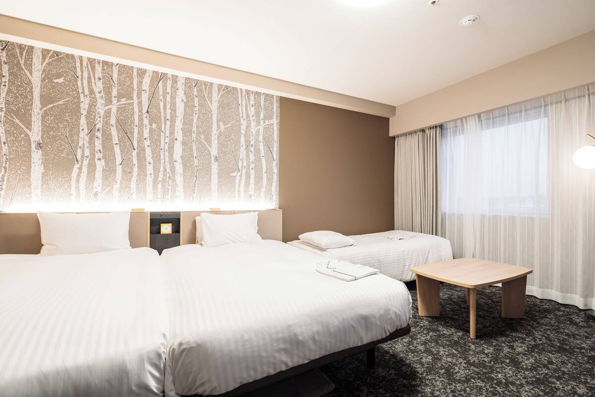 A hotel room with two beds, a small wooden table, and a large window with sheer curtains.