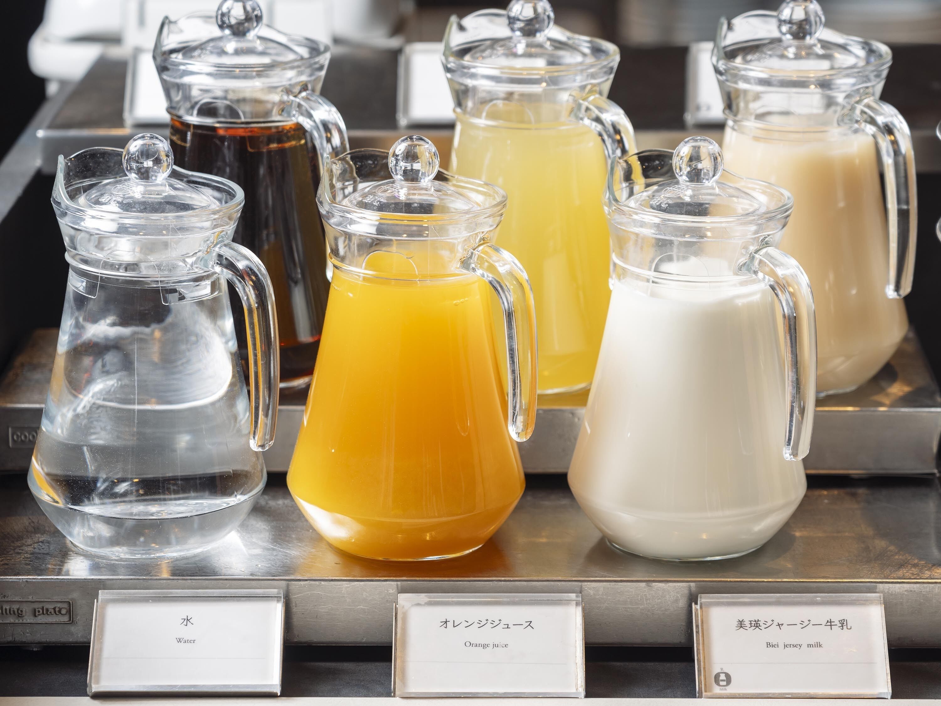 Una estación de bebidas con dispensadores de vidrio transparente etiquetados para agua, zumo de naranja y leche Jersey Biei.