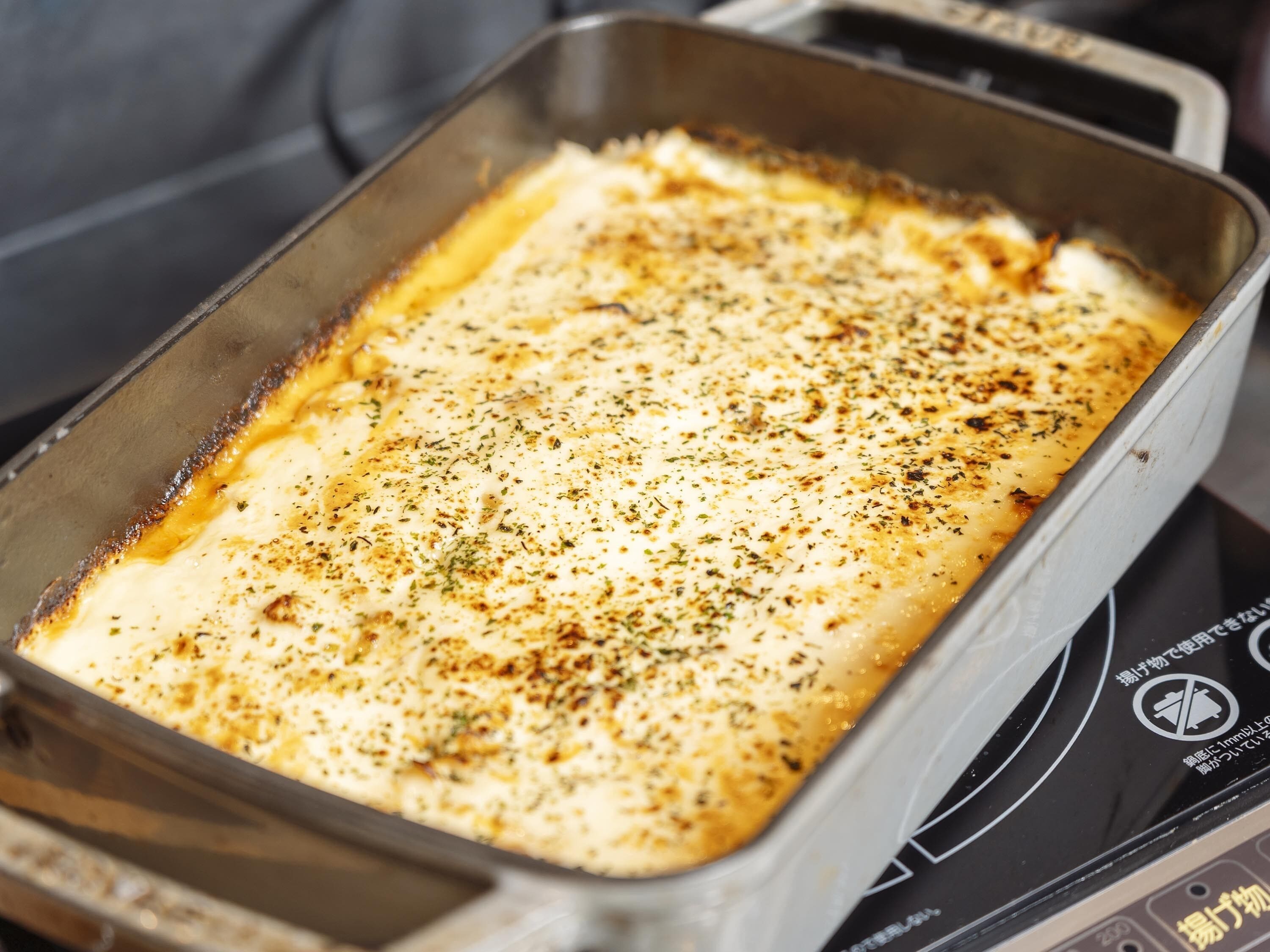 Una fuente metálica rectangular con un plato al horno en su interior, posiblemente lasaña, sobre una hornilla.