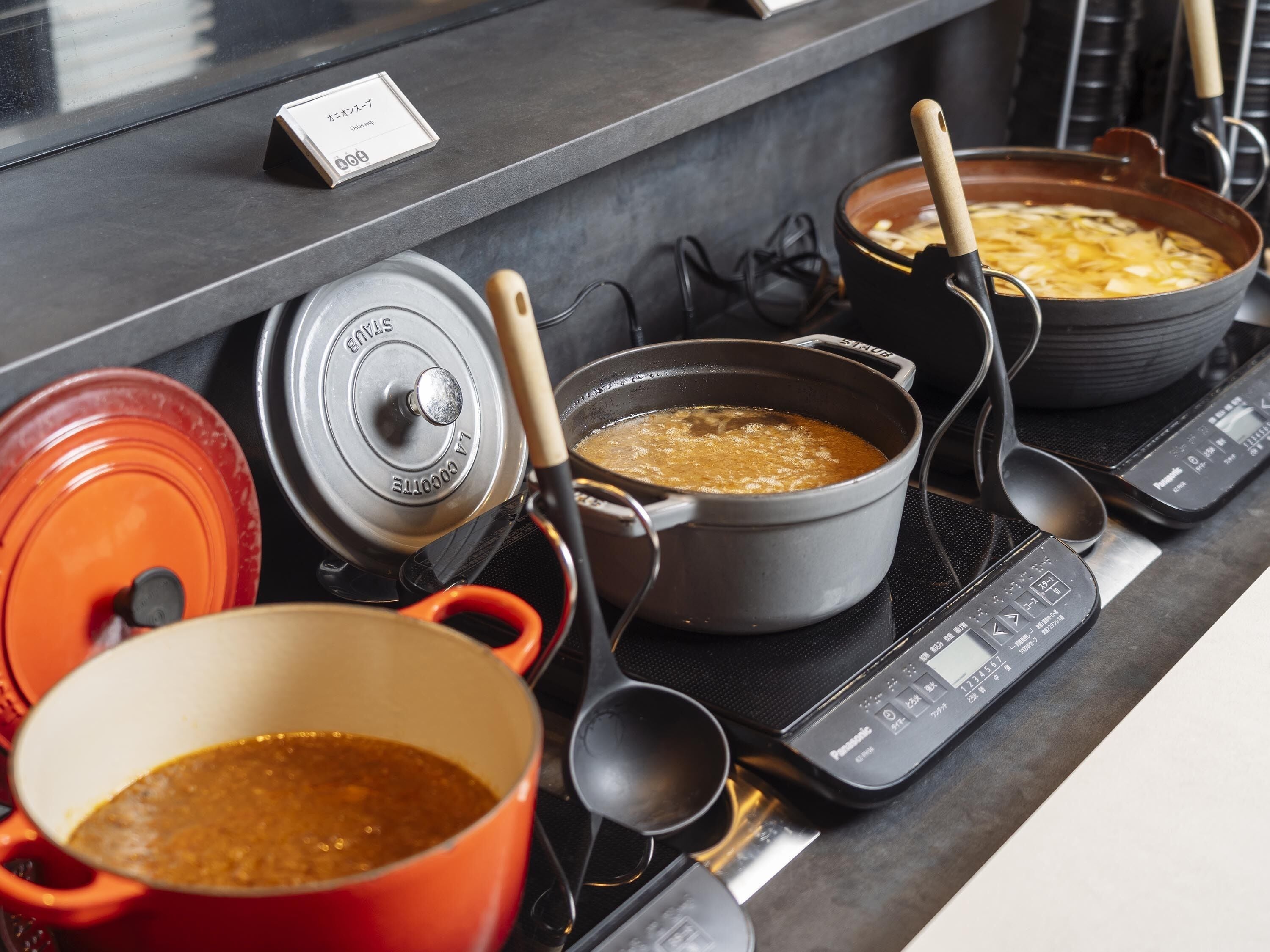 Una cocina con varias ollas y sartenes sobre una estufa, una balanza digital y un cartel que indica un menú.