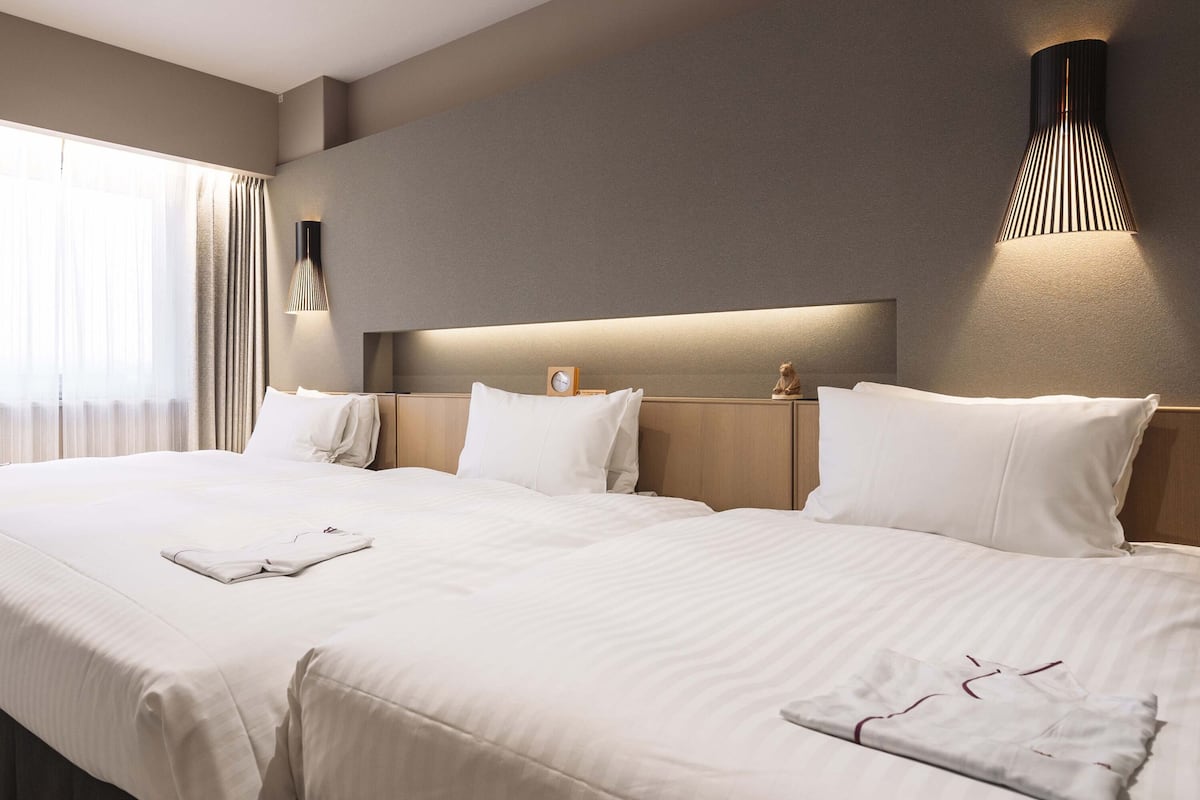 A hotel room with two beds, a bedside table, a clock, and a wall-mounted light fixture.