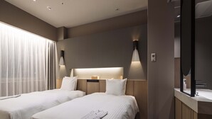 A hotel room with two beds, a large window with sheer curtains, a wall-mounted lamp, and a mirror above the sink.