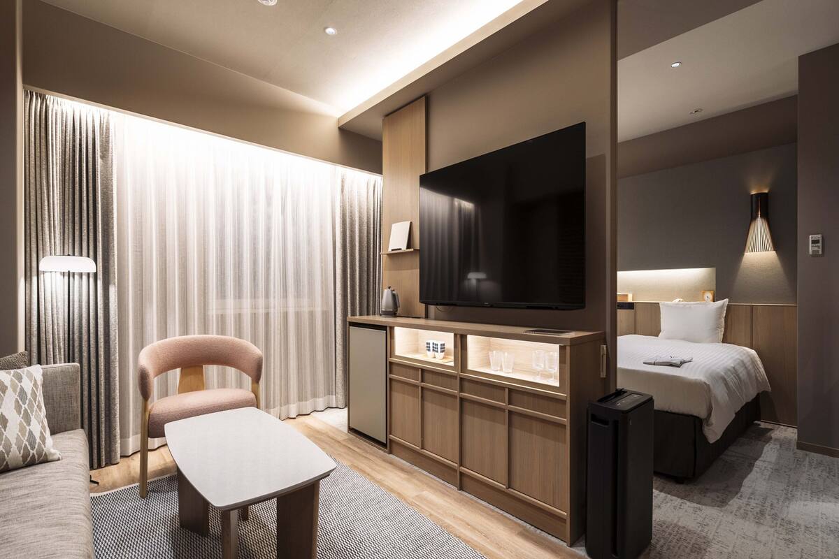 A modern hotel room with a flat-screen TV, a bed, a desk with a chair, and a sofa.