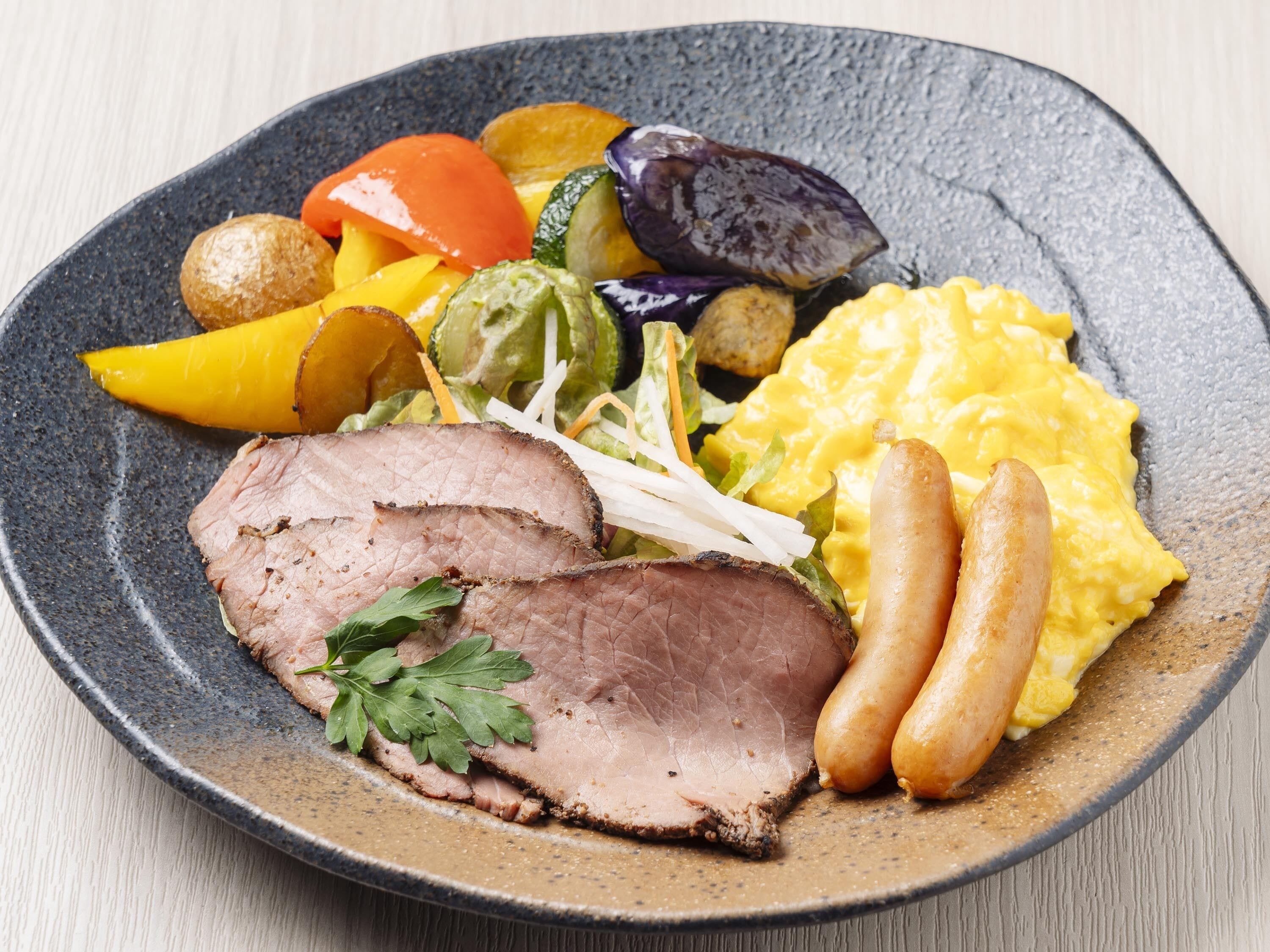 A plate with sliced roast beef, sausages, scrambled eggs, and a variety of vegetables.