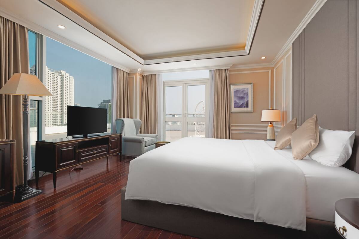 Habitación de hotel con una cama grande, televisión, escritorio con lámpara y vista a edificios altos.