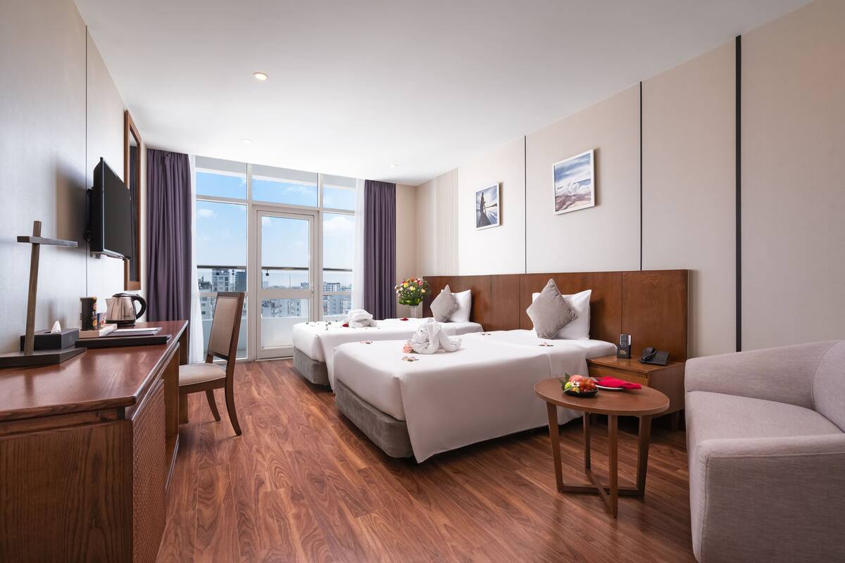 Habitación de hotel con dos camas, un escritorio, un televisor, vistas a la ciudad y un balcón.