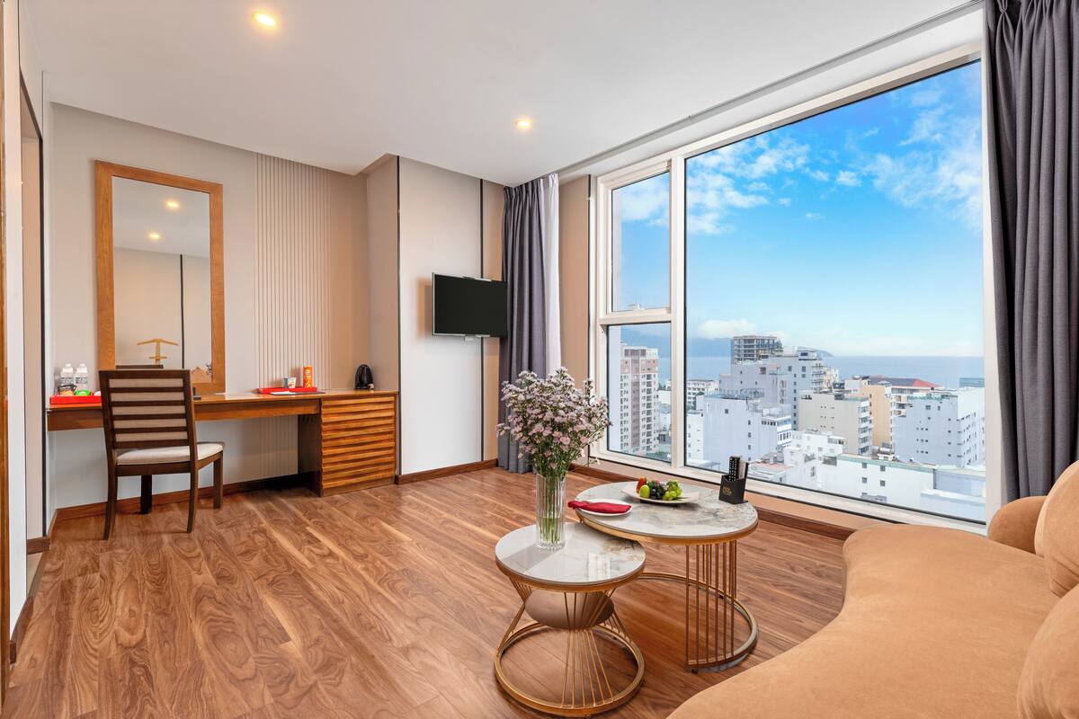 Una habitación moderna con piso de madera, un escritorio con una silla, un televisor, una mesa de comedor con un florero y un ventanal con vista a la ciudad.